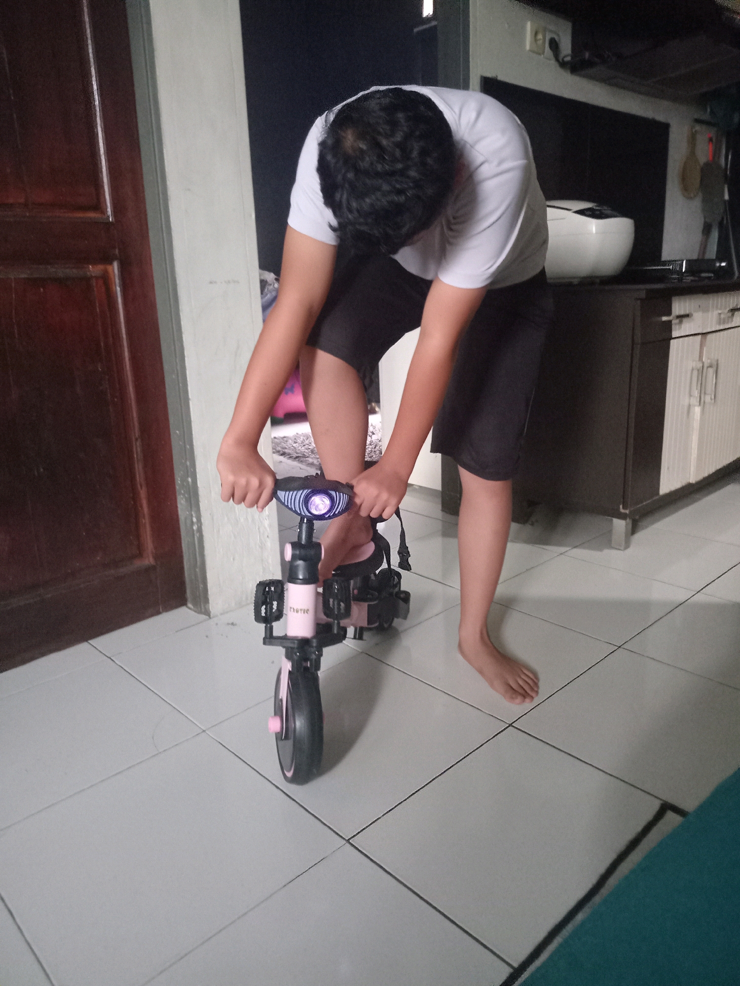 A person with dark hair, wearing a white t-shirt and dark shorts, is bending over to adjust a pink and black children's tricycle. The tricycle has three wheels, with the front wheel featuring a light and pedals. The person's hands are on the handlebars. They are barefoot. The scene appears to be indoors, with tiled flooring and a wooden door visible to the left. In the background, a kitchen counter with appliances and cabinets can be seen. The location is identified as Namo Tualang, Indonesia.
