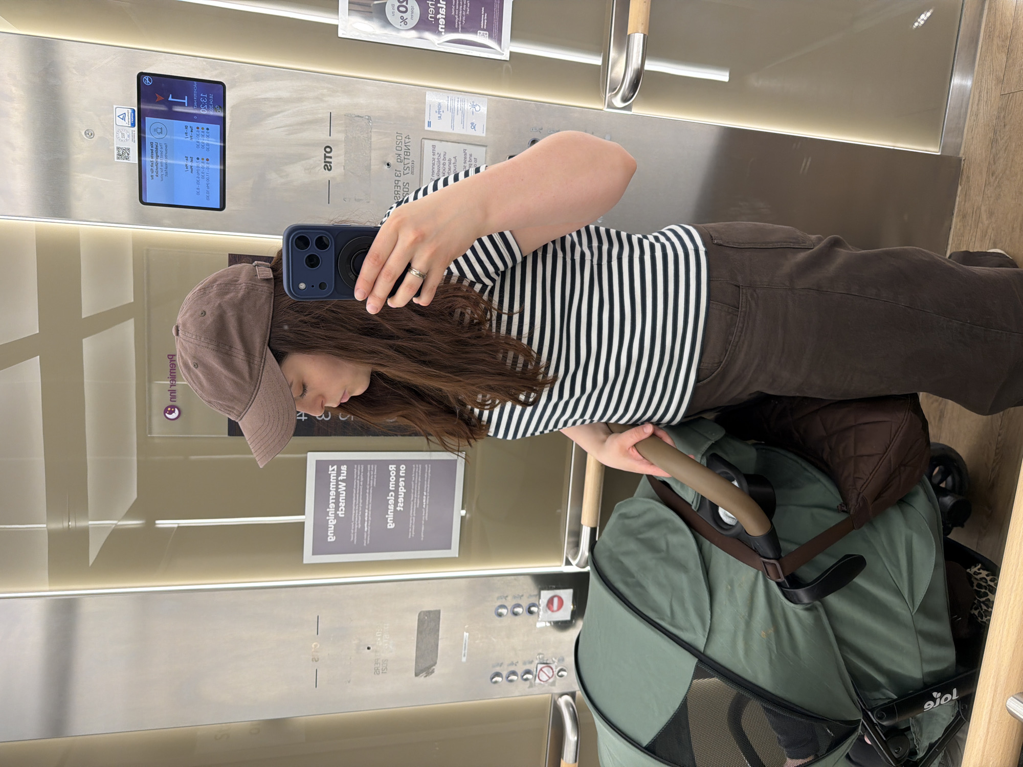 A woman wearing a striped shirt, brown pants, and a brown baseball cap is taking a selfie in an elevator. She is holding a blue smartphone. A green and brown stroller is next to her. The elevator has signage in German, including “JAMES SUSI,” “prinses moo,” and “gnupinistrammis.” There is a digital display showing the time 13:50 and information about the elevator. The elevator is located in the city of Wiesbaden, Germany.