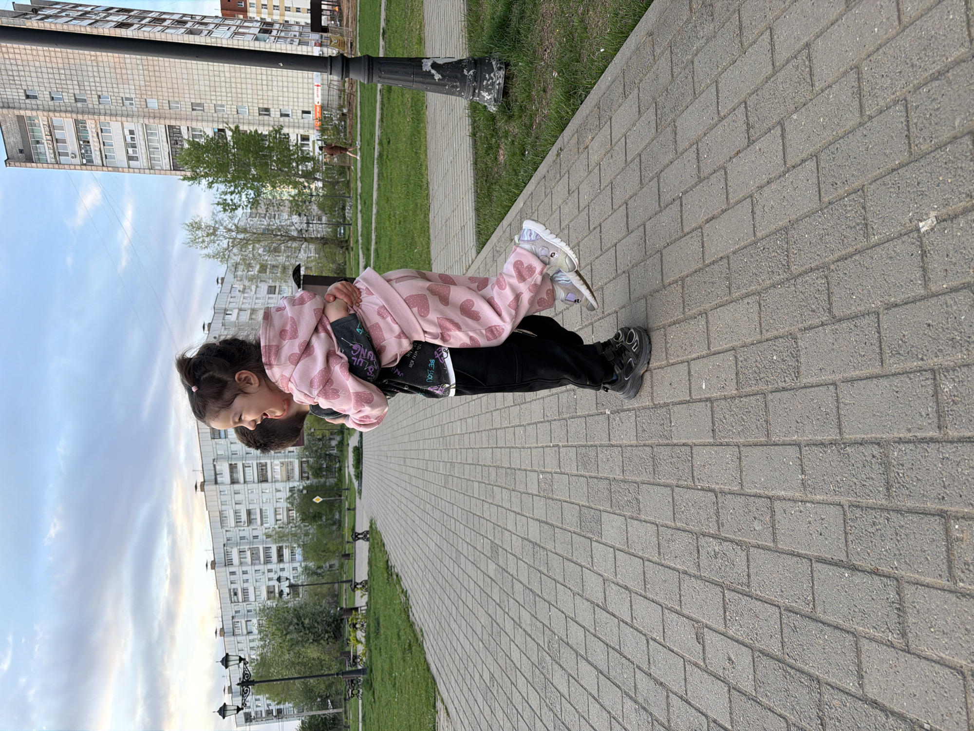A young girl in a pink heart-patterned outfit is being carried on the back of a person wearing black pants and shoes. They are on a brick walkway lined with grass and lamp posts. Buildings are visible in the background. The location is likely a park in the city of Chaikovsky, Russia.