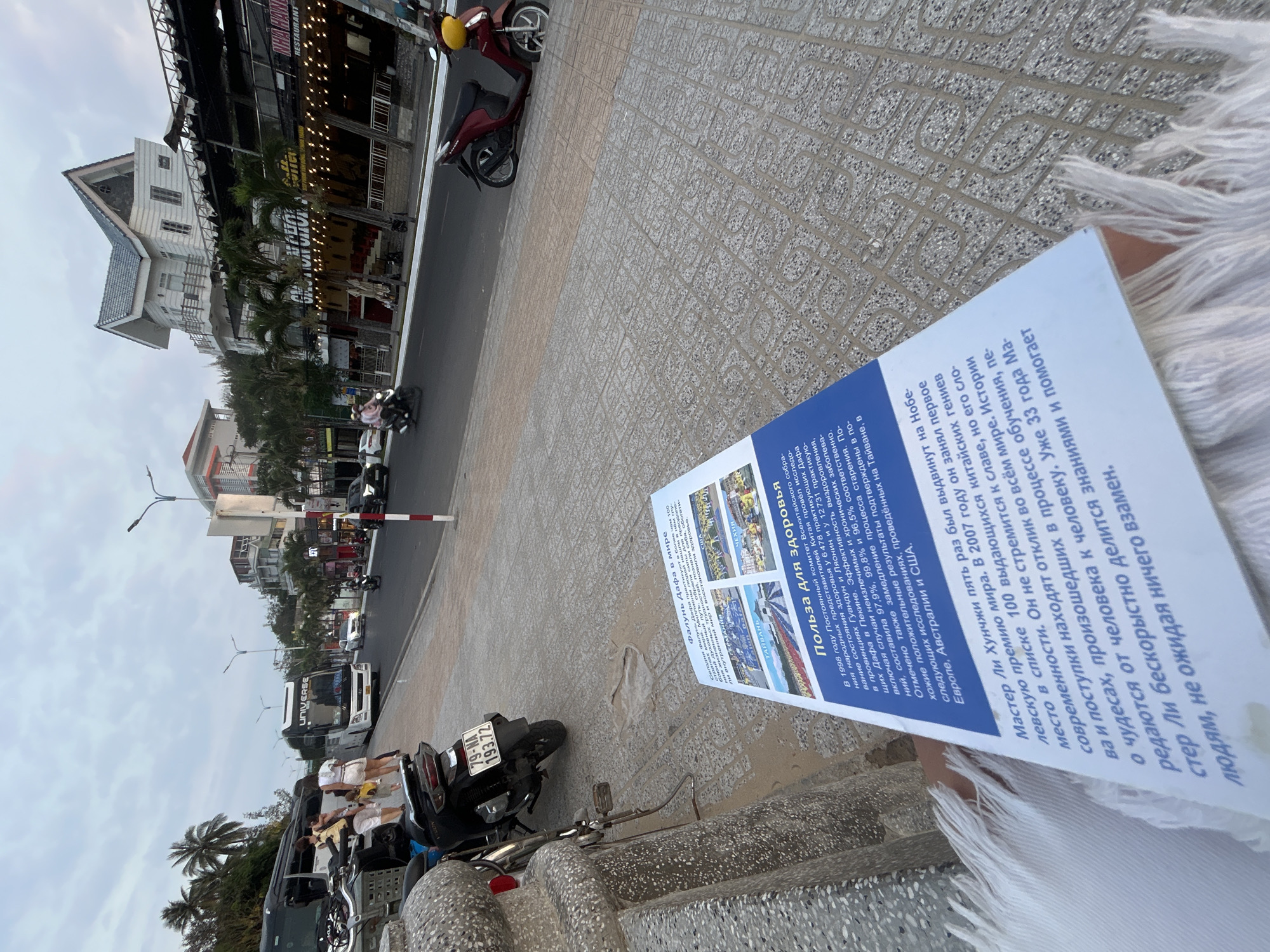 A person holds a brochure while standing on a tiled sidewalk in Bắc Nha Trang, Vietnam. The brochure promotes "Health Benefits" and is printed in Russian. In the background, a street features motorbikes, cars, and a bus. Buildings line the street, with some having lights and restaurant signage. Palm trees are visible along the street. The sky is overcast.