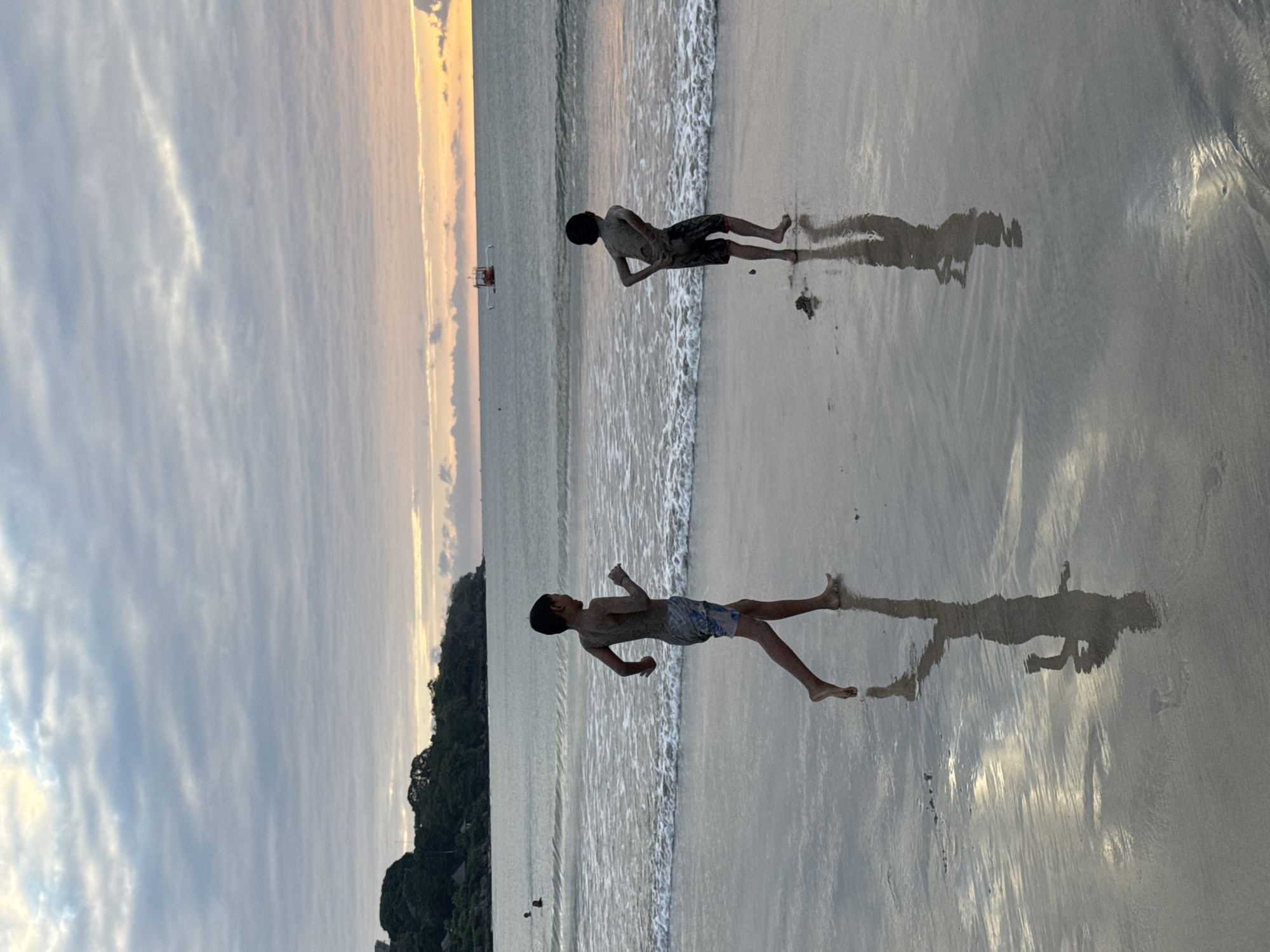 Two young boys are running on a wet sandy beach in Kuta Selatan, Indonesia, near the water's edge as a boat is visible in the distance on the ocean. The sky is filled with clouds, transitioning into a warm sunset glow on the horizon. The boys are reflecting in the wet sand, creating mirrored images of their silhouettes. The boy on the left is wearing patterned swim trunks, and the boy on the right is wearing dark swim trunks. The waves are gently washing onto the shore. The environment appears to be a tropical beach with lush green vegetation visible on a hillside in the background.