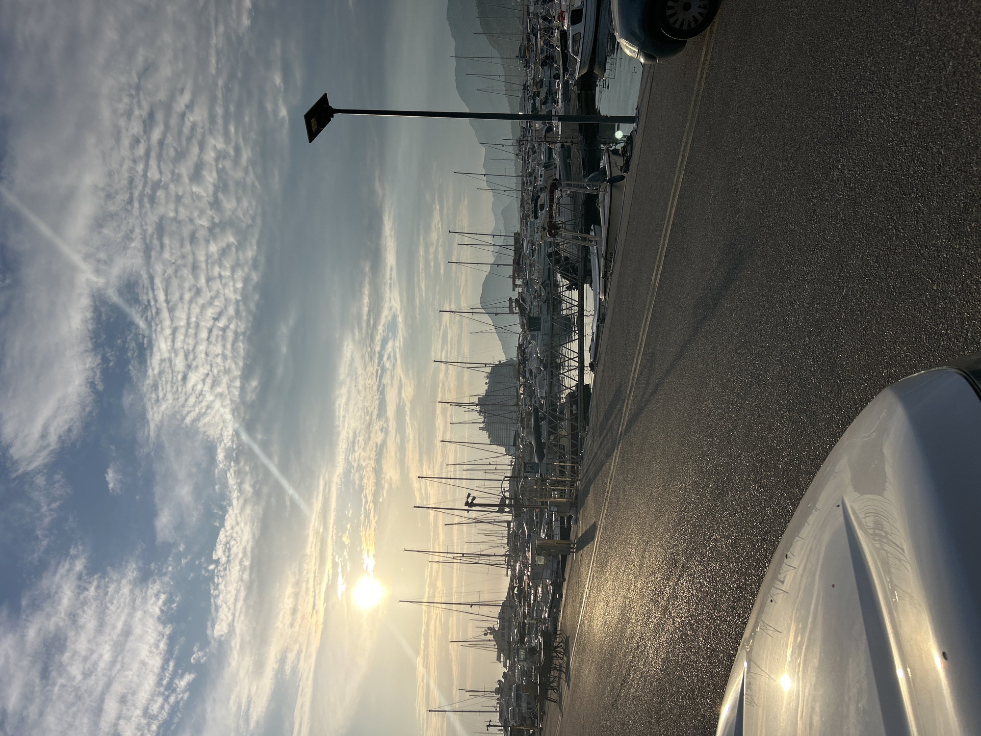 The image depicts a marina with numerous sailboats and yachts docked side-by-side. The scene is set in Bar, Montenegro. The sun is low in the sky, casting a warm glow and long shadows, suggesting it is either early morning or late afternoon. The sky is partly cloudy, with wispy clouds scattered across a pale blue expanse. In the background, mountains rise, adding to the scenic backdrop of the location. A paved road runs alongside the marina, with double yellow lines indicating a traffic lane. The wet appearance of the road suggests recent rain or damp conditions. A modern street lamp with a solar panel stands tall near the water's edge. The foreground shows the hood of a white car, reflecting the sunlight and the surrounding environment. The overall mood is serene and tranquil, capturing the quiet beauty of a marina at a peaceful time of day. There is no visible text in the image.
