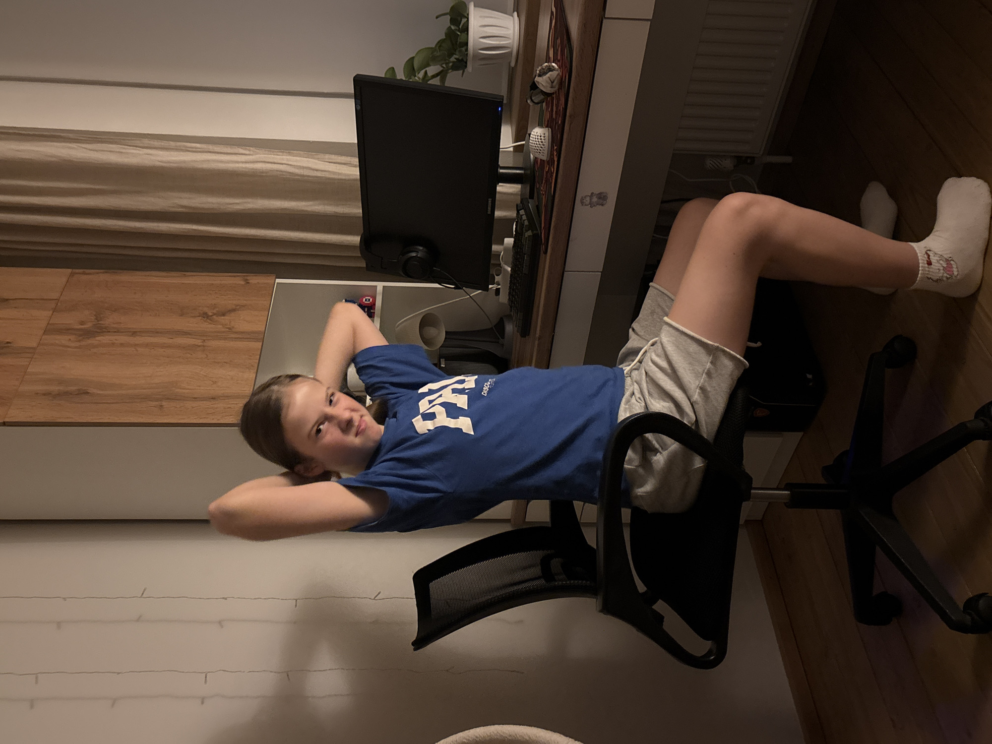 A young person is reclining in an office chair in an indoor setting. The person is wearing a blue t-shirt, grey shorts, and white socks. They are positioned in front of a desk with a computer monitor, keyboard, and mouse. There is also a plant on the desk. The setting appears to be a home office or bedroom. The location in Chaikovsky, Russia is not apparent.