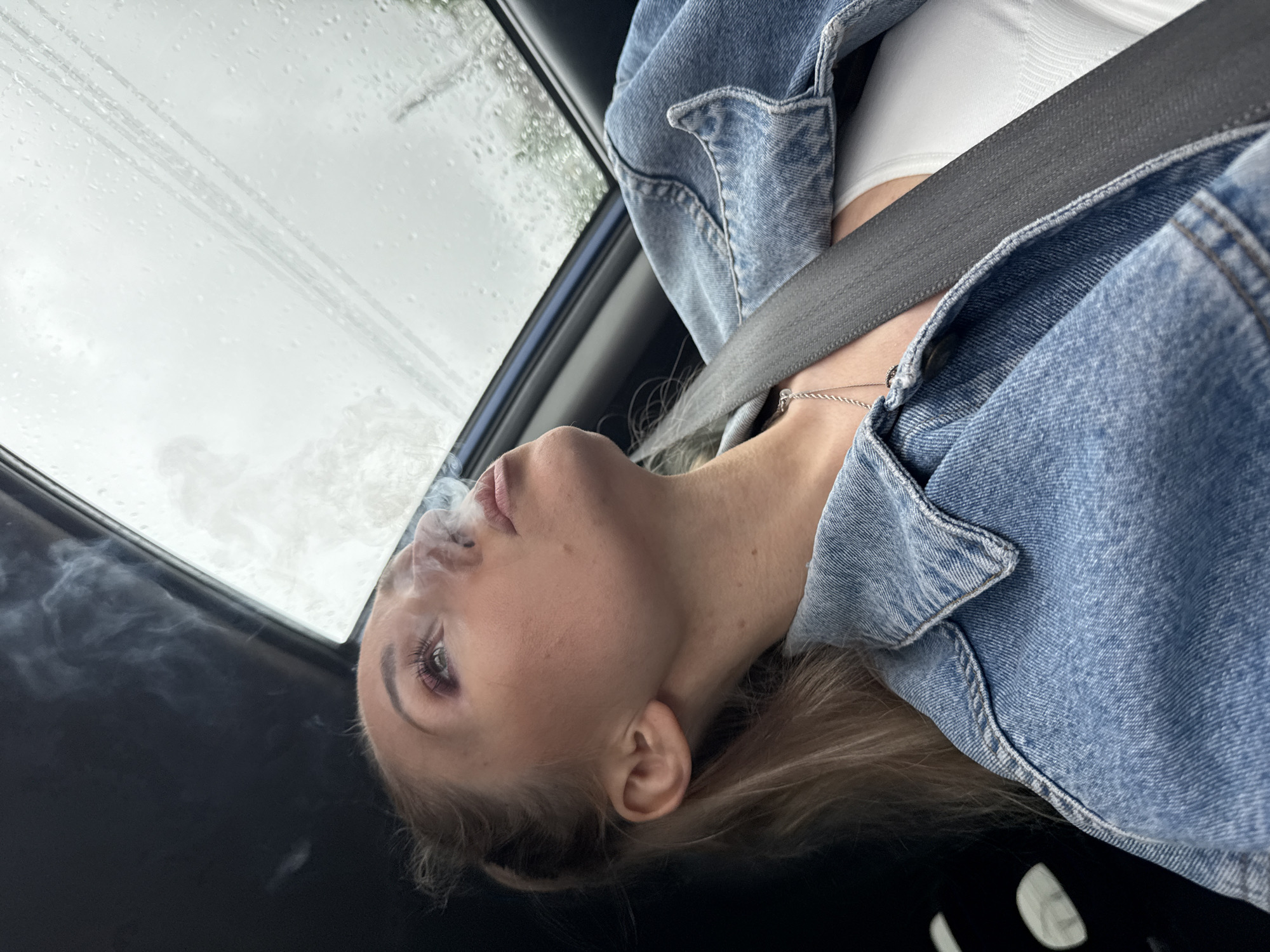 A young woman is seated in a car, wearing a white top and a blue denim jacket with a seatbelt across her chest. She is exhaling smoke. The sky is visible through the car's roof window and appears overcast with rain. There is no indication of a location in the city of Chaikovsky, Russia.