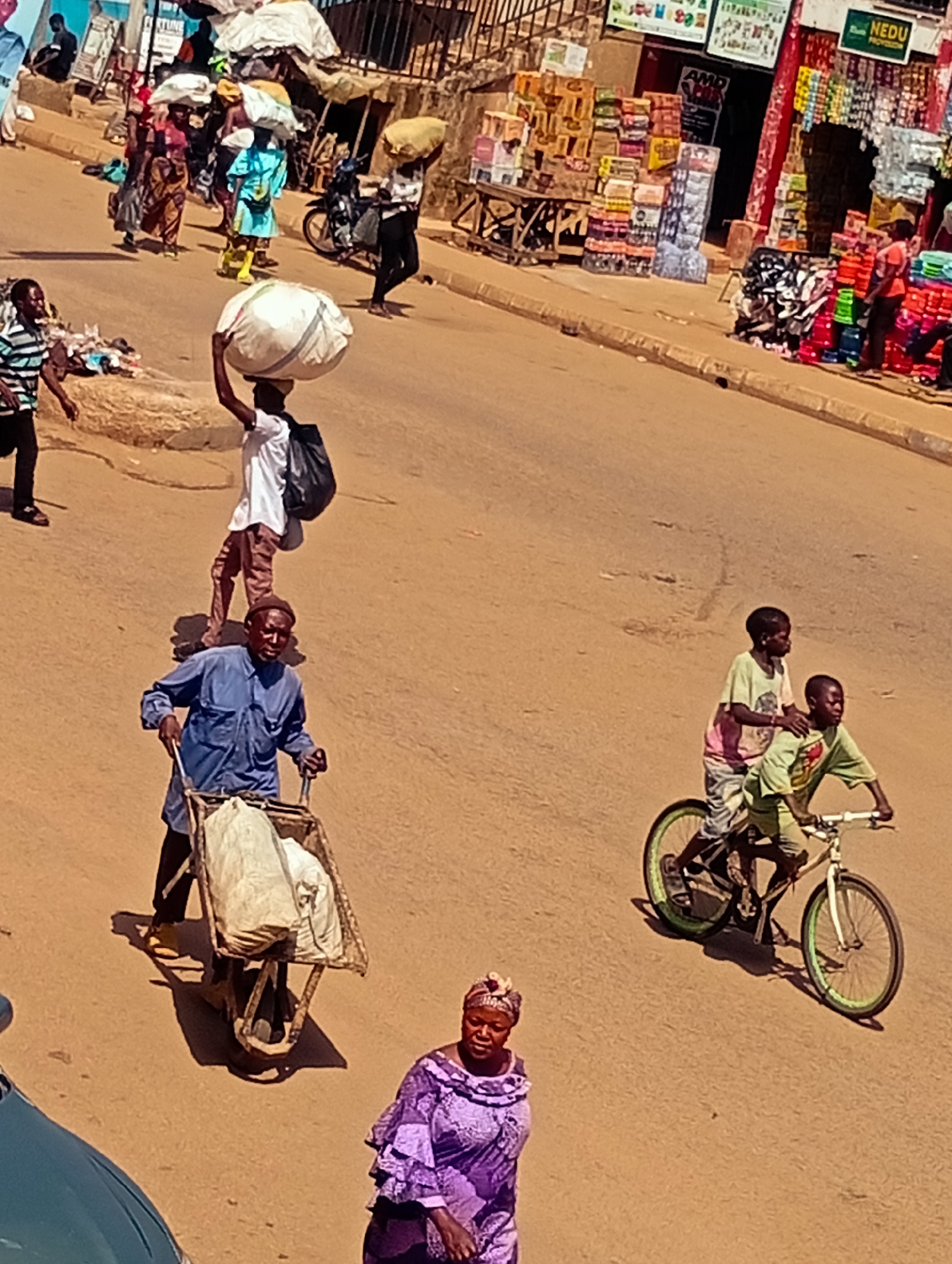 In Bukuru, Nigeria, a bustling street scene unfolds under daylight. A man wearing a blue shirt and a dark cap pushes a wheelbarrow filled with large, white sacks along a dusty road. To his left, a person in a white shirt and brown pants carries a large, round, white object on their head. Further up the street, several individuals are seen carrying bundles on their heads, navigating through the crowd. On the right side of the road, two young boys are riding a bicycle, one pedaling and the other sitting behind him. In the foreground, a woman in a vibrant purple dress and headscarf walks towards the viewer. The background reveals shops with colorful displays of goods, suggesting a market or commercial area. The overall environment appears to be an outdoor marketplace characterized by pedestrian traffic and commercial activity.
