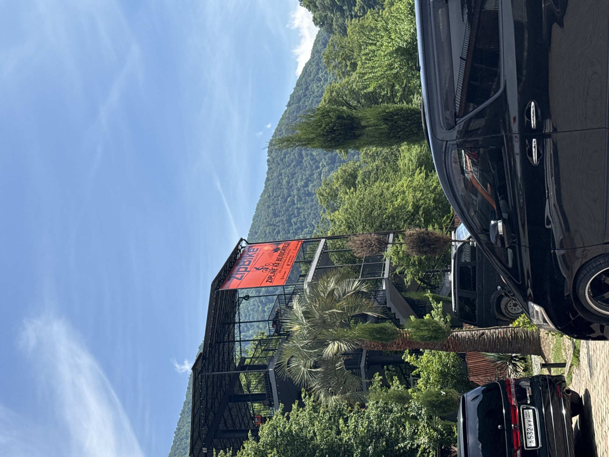 The image depicts an outdoor scene in Bzypta, Abkhazia, under a clear blue sky with sparse white clouds. A multi-level, open-air structure, likely commercial, stands centrally, constructed with a dark metal frame and glass panels, featuring exterior staircases and balconies. A large red banner on the structure displays "ZipBike - Абхазия" and "ZIPLINE НА ВЕЛОСИПЕДЕ" (Zipline on a bicycle) along with a graphic of a cyclist on a zipline.
Lush green foliage surrounds the structure, including dense trees covering mountains in the background. A prominent palm tree is positioned in front of the structure, with several slender coniferous trees also visible.
Three black vehicles are present in the foreground and mid-ground. On the far left, a black minivan or large SUV is parked, displaying an Abkhazian license plate "A 532 YY" with "ABM." Partially cropped on the right, another black minivan or large SUV with custom wheels is parked, showing its side profile. A third black SUV, possibly a UAZ Patriot, is visible in the mid-ground behind the palm tree and the left-most minivan. The ground in the parking area is paved. No individuals are visible.