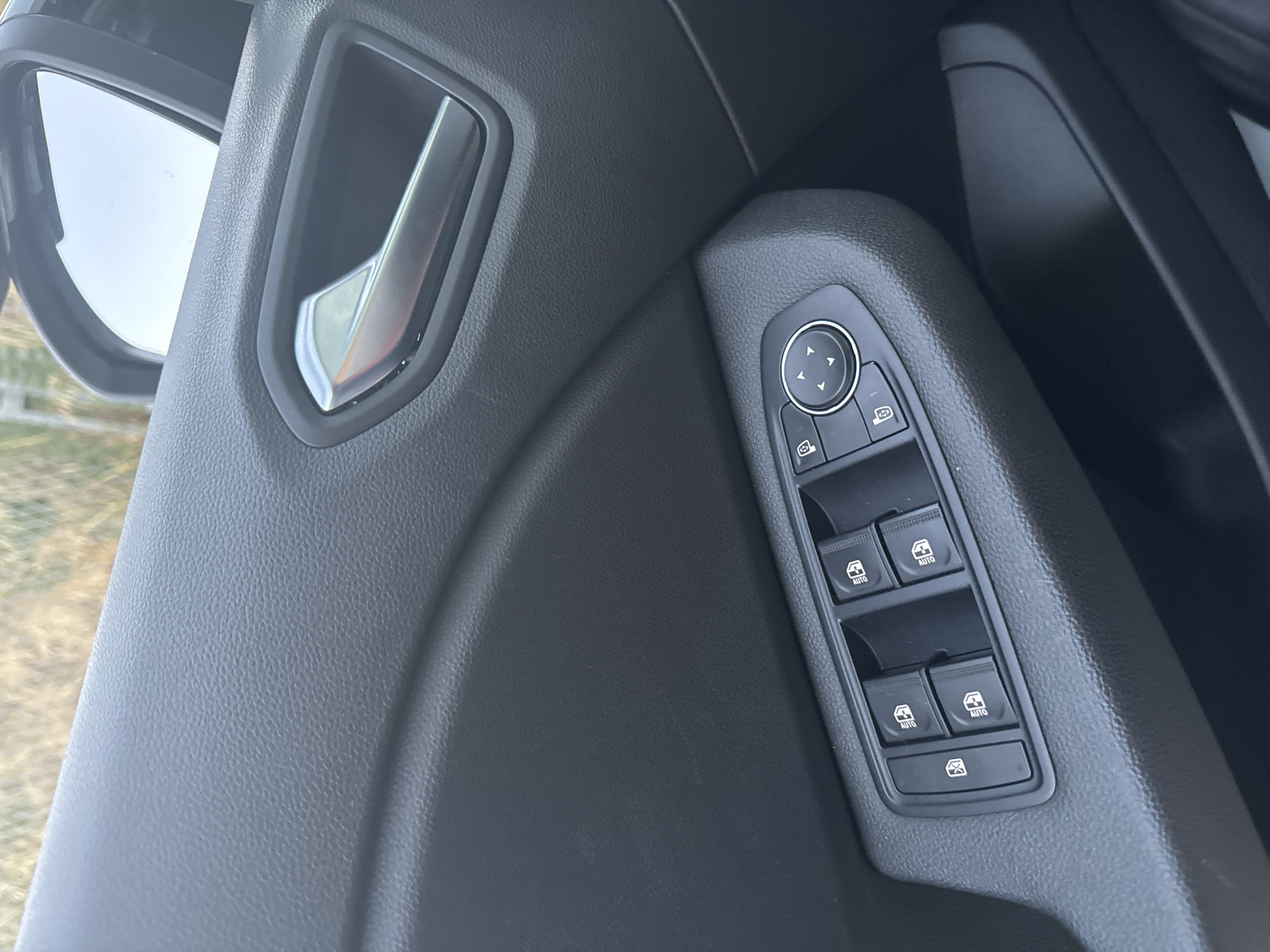 The image shows a close-up view of the interior of a car door, specifically focusing on the window controls and door handle. The car appears to be parked outdoors, with a blurred background that suggests foliage or a grassy area, possibly indicating a location in Murcia, Spain, given its Mediterranean climate and abundant greenery. The car's interior is primarily black and grey, with a textured finish on the door panel. The window control panel features several buttons, including those for controlling the windows and side mirrors, indicated by icons of car windows and directional arrows. A silver door handle is visible above the window controls. No people or actions are depicted in the image; it is a static shot of the car's interior.