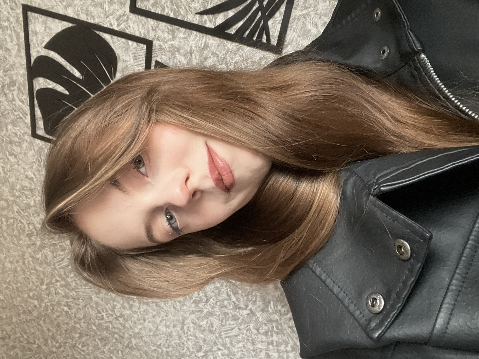 A close-up portrait, taken indoors in Фоки, Russia, features a young woman with long, straight, light brown hair. Her fair skin contrasts with her light-colored eyes, which are open and look directly at the viewer. She is wearing a black leather-like jacket, details of which, including a metallic zipper and snaps, are visible on her right side. Her lips are adorned with a muted pinkish-brown lipstick, and she has subtle eye makeup, including eyeliner and mascara. Her expression is subdued, with a slight, enigmatic smirk. The background is a light-colored, textured surface, possibly a rug or wallpaper, on which she appears to be resting. Behind her head, two black, framed, stylized leaf decorations are visible on the wall. The overall image is rotated 90 degrees clockwise. There is no visible text.
