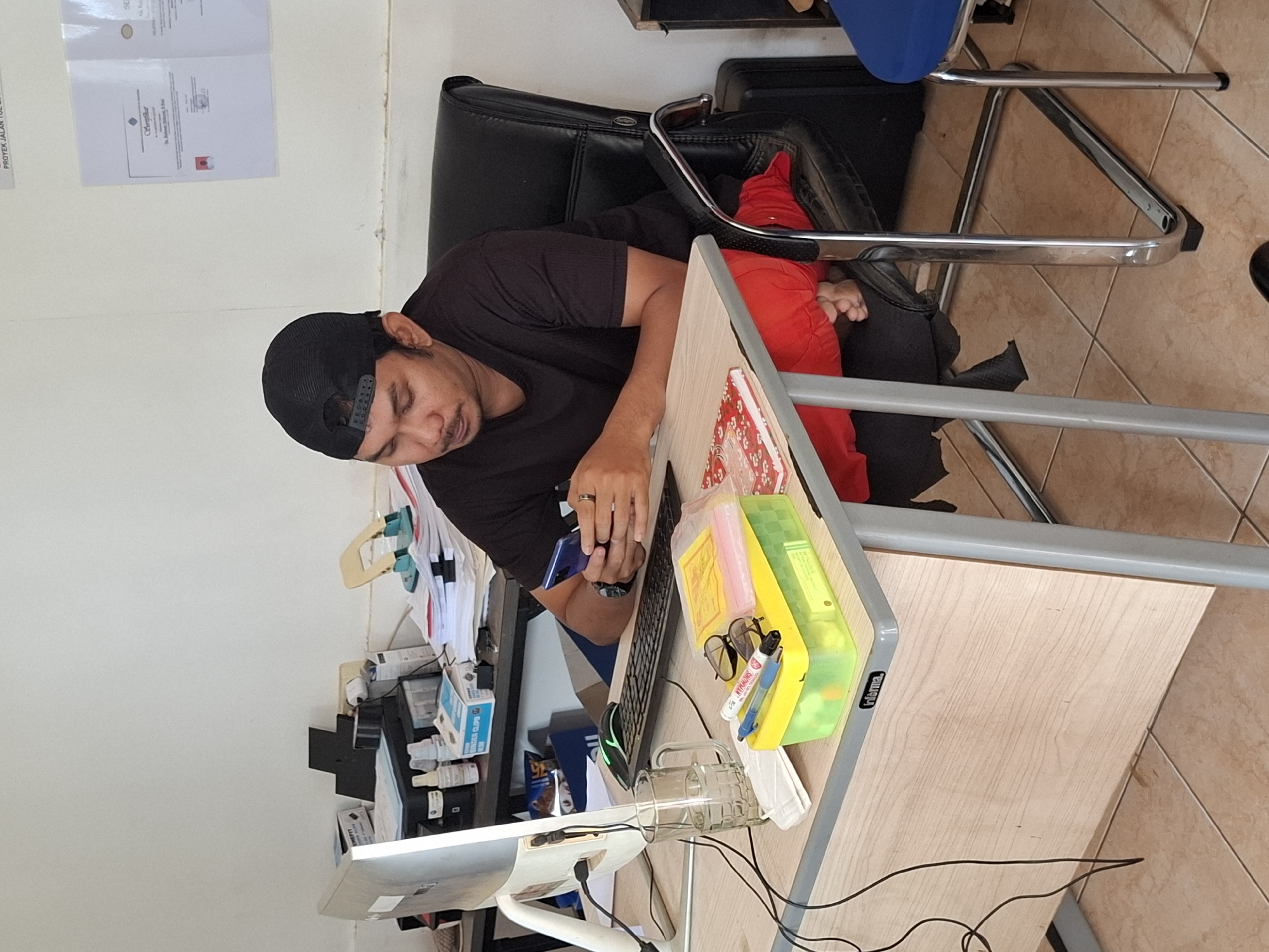 In an indoor office setting located in Ciheulang Tonggoh, Indonesia, a man wearing a black t-shirt and a black baseball cap is seated at a light-colored desk. He is holding a smartphone in his hands and appears to be focused on it. A computer monitor, keyboard, and mouse are visible on the desk, along with a glass of water, a yellow storage container filled with stationery items, and a notebook. The man is dressed in red pants and is sitting in a black leather office chair. The surrounding environment includes office equipment and documents.