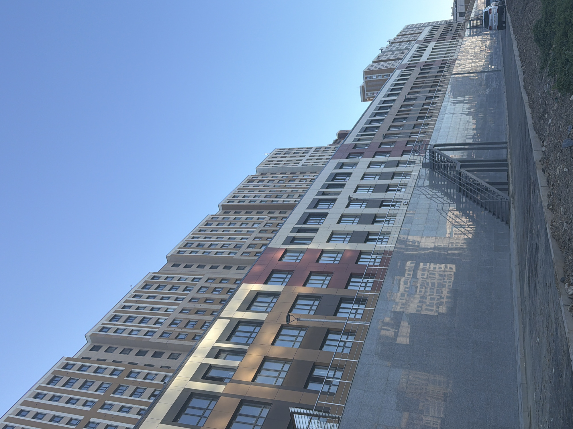 The image shows a multi-story residential building with a facade featuring a combination of beige, brown, dark red, and white panels, each section housing multiple windows. The building is situated on a tiered structure, with a large, reflective gray stone wall forming the base. A metal staircase is visible ascending along this wall. The sky is clear and blue. In the foreground, there is a paved area and a dirt embankment with some sparse vegetation. Two cars are parked to the right of the building. The architectural style and the clear blue sky suggest a modern development, likely in the city of Astana, Kazakhstan, given the context provided.