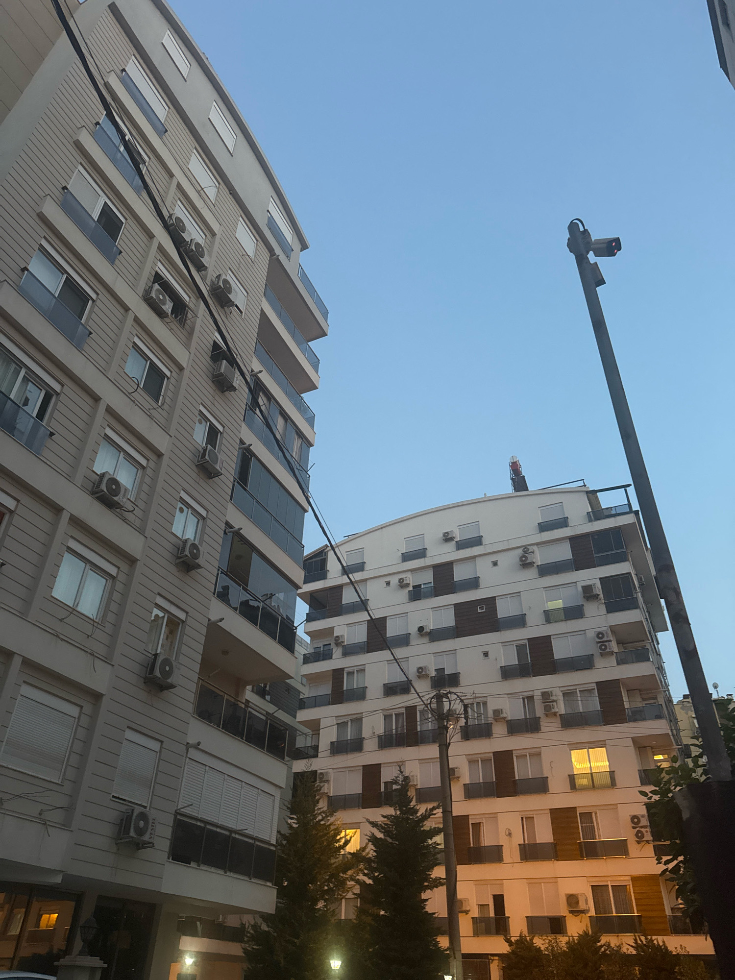 Two multi-story apartment buildings are pictured in Antalya, Turkey, on a clear day. The building on the left is beige with horizontal siding and numerous windows, many with air conditioning units mounted below them. Balconies with glass railings are visible on several floors. The building on the right is white with brown accents around some windows and also features multiple air conditioning units. A traffic light with a red signal is mounted on a tall pole to the right of the frame, obscuring part of the sky. Electrical wires run between the buildings. Trees are visible in the foreground, and lights are illuminated in some of the apartment windows.