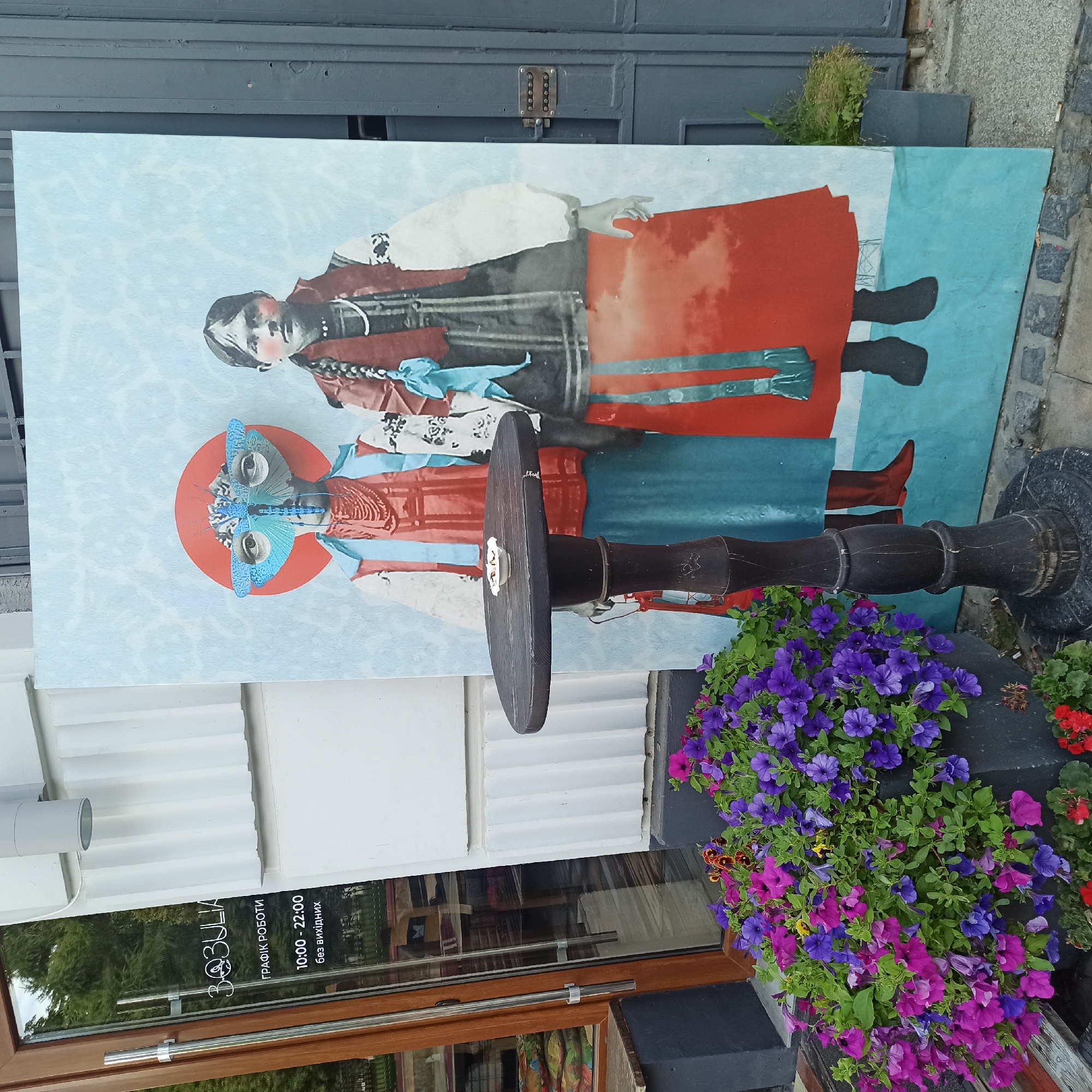 The image depicts a street scene outside a shop in Chernivtsi, Ukraine. On the left, a glass door with wooden framing, bearing the Cyrillic text "ЗОЯ" and "графік роботи 10:00 - 22:00 без вихідних" (meaning "Zoya" and "working hours 10:00 - 22:00 without days off"). A large, colorful mural of two figures, one wearing a mask resembling a dragonfly and the other a traditional Ukrainian outfit, stands prominently in the center. In front of the mural, a dark, antique-style wooden table with a small white dish on top is positioned. To the left of the table, a large planter overflows with vibrant purple and pink petunias. The background features a white textured wall and a grey metal gate. The ground is paved with cobblestones and some concrete.