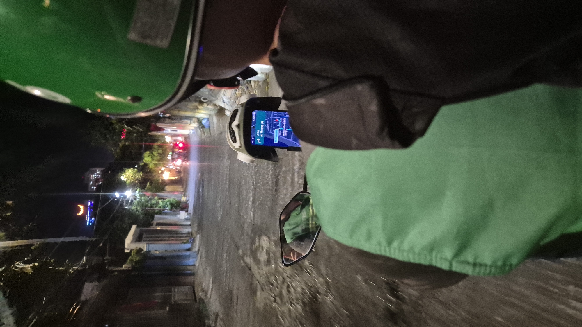 A first-person perspective shot shows a person riding a motorbike at night in Da Nang, Vietnam. The rider is wearing a green helmet and a black shirt, with a bright green bag on their back. In front of them, a smartphone mounted on the handlebars displays a navigation app with directions to "An Thượng 30". The screen shows a route and estimated travel time. The motorbike is on a narrow street lined with buildings. In the distance, streetlights and illuminated buildings create a warm glow. A prominent building with a crescent moon-shaped light fixture is visible on a hillside in the background. The wet pavement suggests it might have rained recently. The overall scene captures a typical urban night in Da Nang, with a focus on the practicalities of navigation for a motorbike rider.