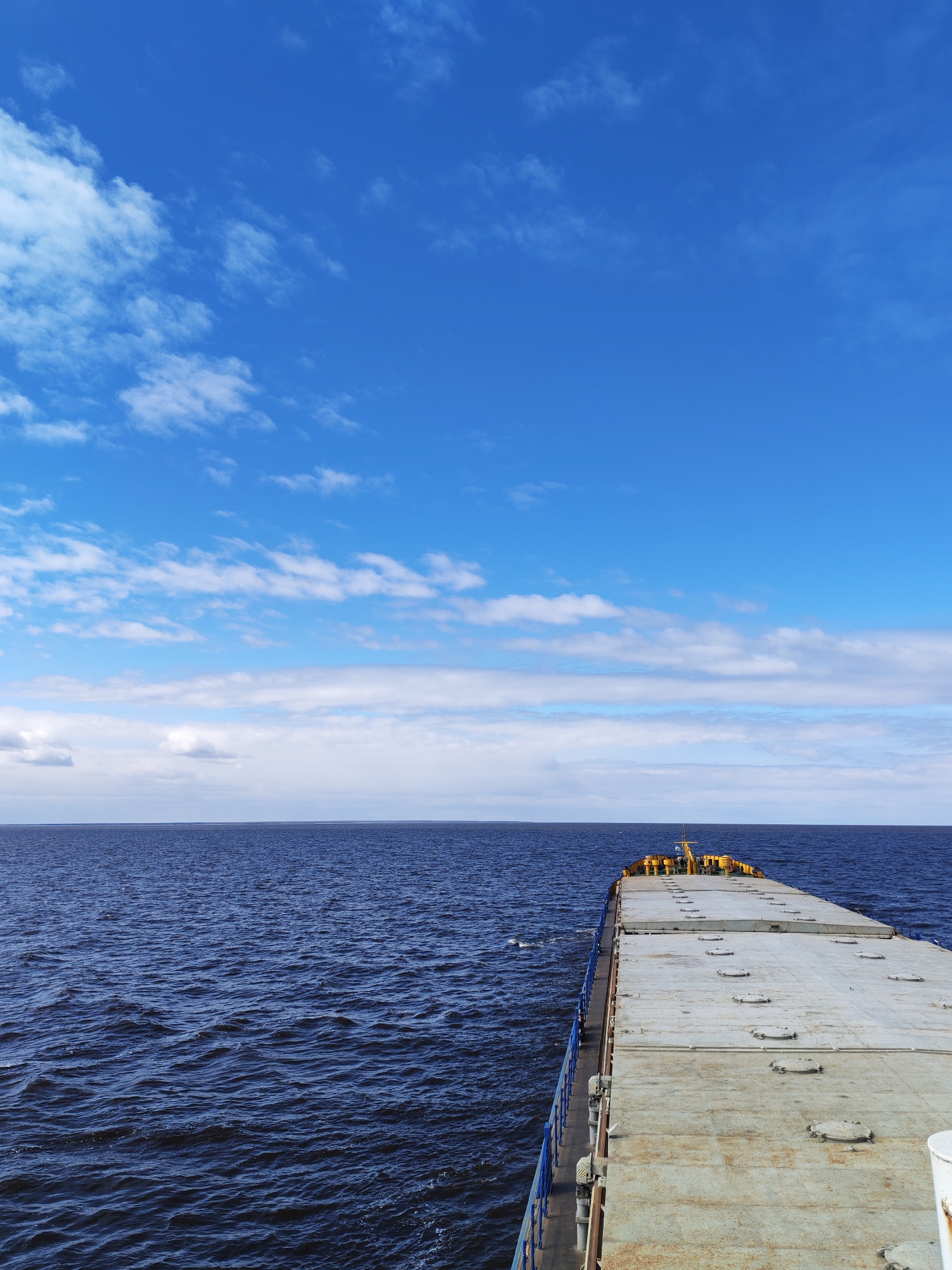 The image presents a forward-looking view from the deck of a ship on a body of water, likely a large lake or sea. The sky occupies the upper portion of the frame, displaying a bright blue expanse punctuated by scattered white clouds. A distinct horizon line separates the sky from the dark blue, slightly choppy water below. The ship's deck, a flat, gray surface with multiple access hatches and a blue railing along the side, extends toward the horizon. The perspective indicates the camera is positioned near the front of the ship, looking toward the bow. There are hints of yellow-colored equipment towards the ship's bow. No people are visible in the frame. The location is unlikely to be Pochinok, Russia, since Pochinok is not a coastal city or located near any large body of water where such a ship would be navigating.