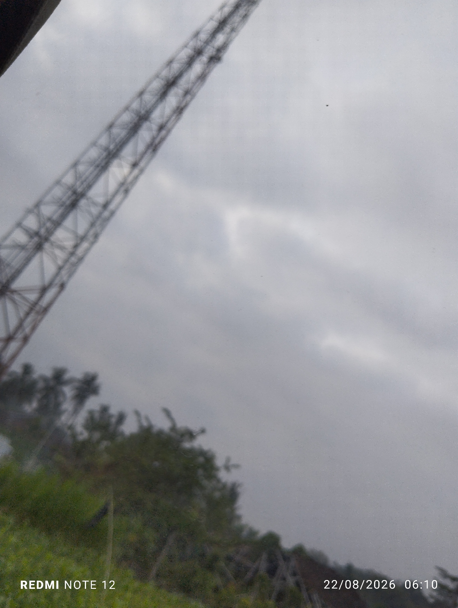 An overcast sky dominates the upper portion of the image, with a tall, dark metal tower structure angled from the upper left corner, extending towards the center. The lower portion of the image shows a blurred view of green foliage and trees, suggesting an outdoor environment. The bottom left corner displays the text "REDMI NOTE 12", and the bottom right corner shows the date and time "22/08/2026 06:10". The location is identified as Dipan, Indonesia.