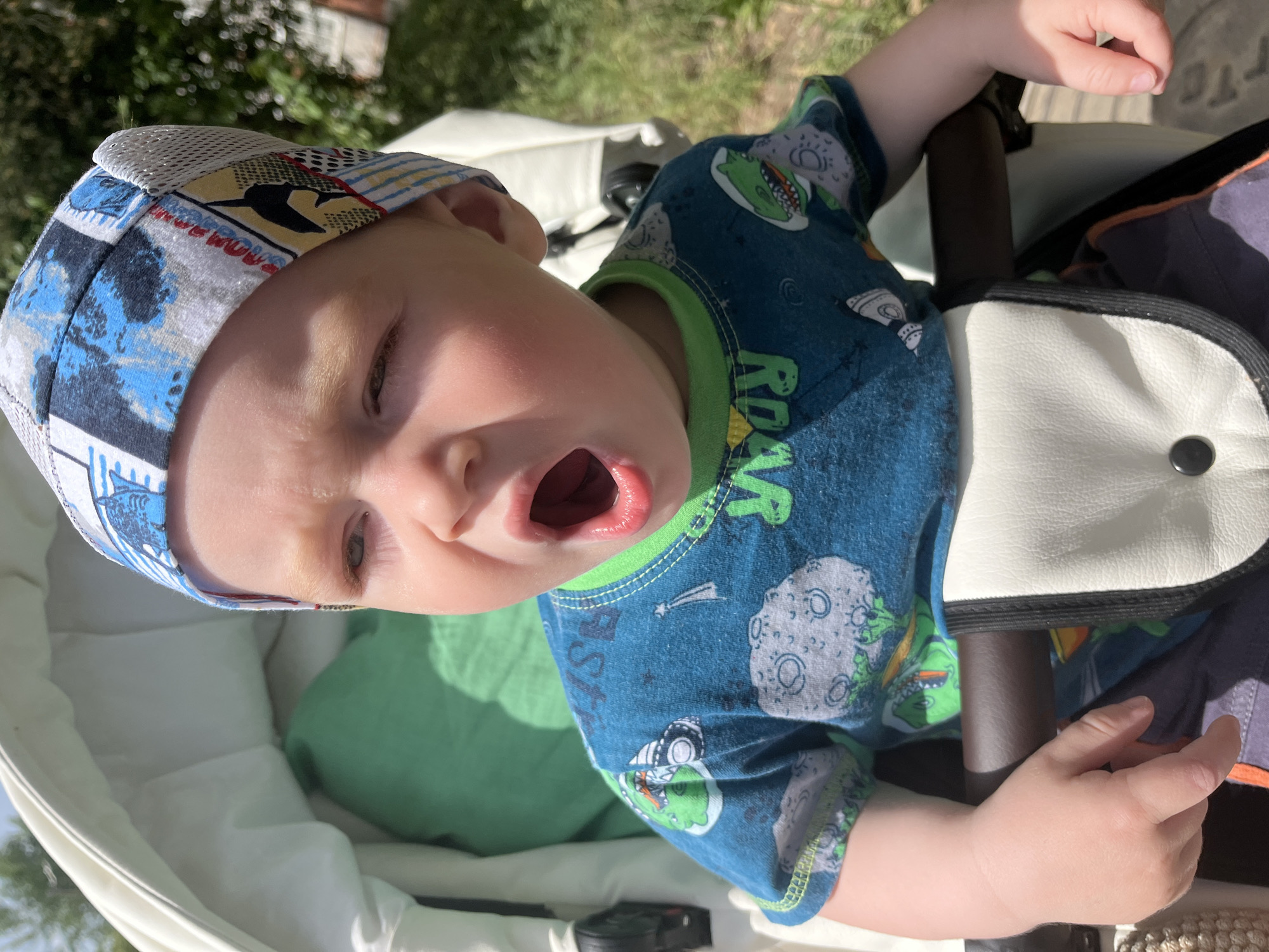 A young child wearing a patterned blue shirt and a cap is sitting in a white stroller outside. The child has their mouth open. A green cushion is also visible inside the stroller. The location cannot be determined.