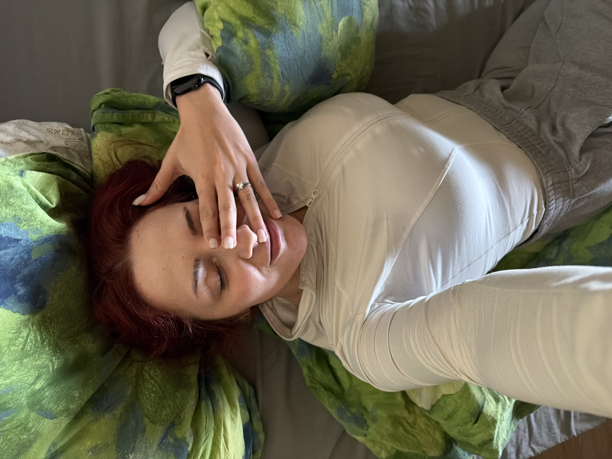 A woman with auburn hair is lying on a bed with her eyes closed. She wears a white zip-up top and gray sweatpants. Her left hand is raised, partially covering her face, and she has a ring on her ring finger. She is lying on a green and blue patterned pillow and blanket. A smartwatch is visible on her left wrist. The bed has gray sheets. The location is not discernible and thus can not be confirmed to be Sofia, Bulgaria.