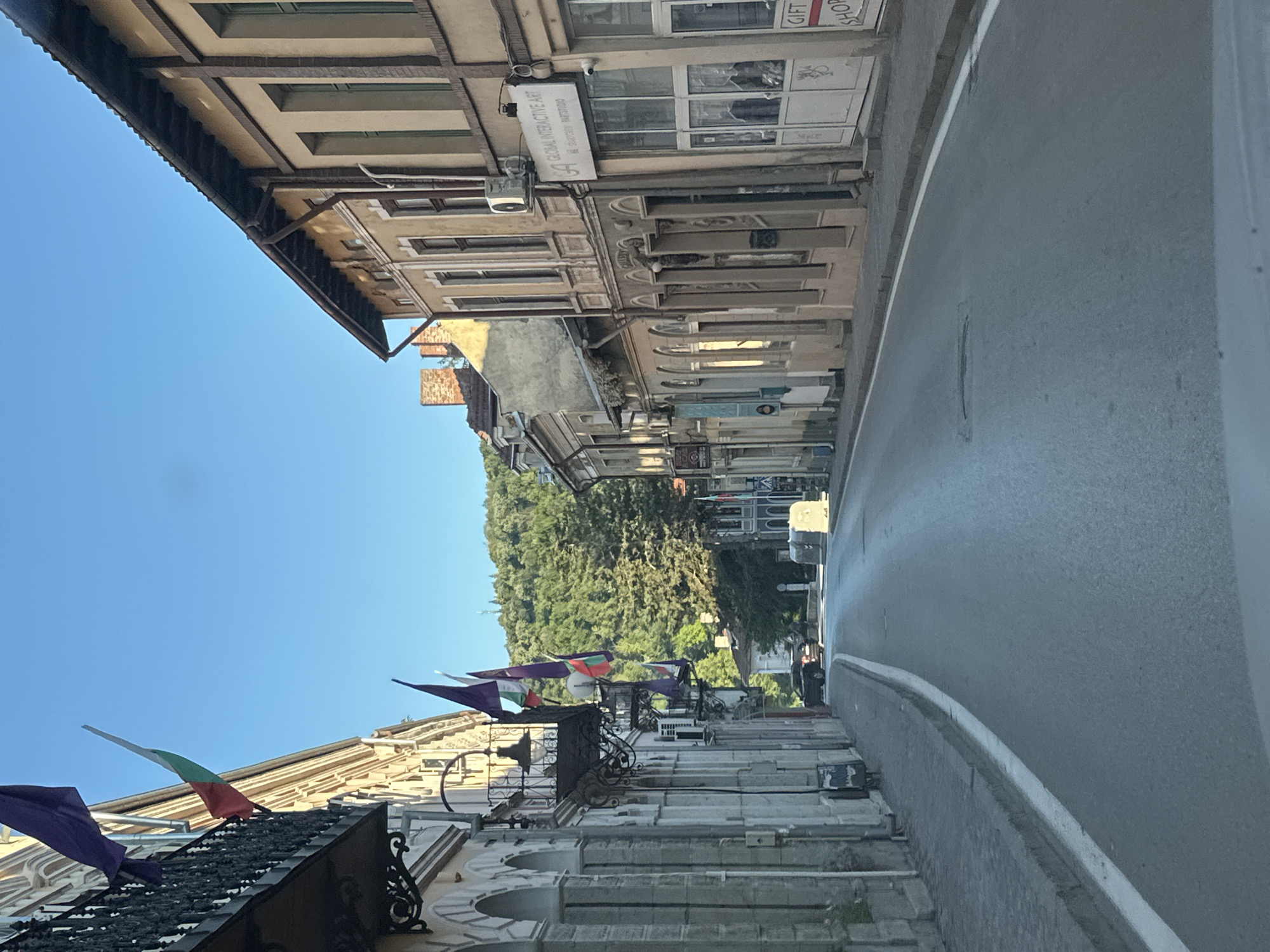 The image captures a street scene in Veliko Tarnovo, Bulgaria. On the left side of the frame are buildings with architectural details such as arched entryways and wrought iron balconies adorned with Bulgarian flags. The buildings have a vintage aesthetic. The street is paved and curves slightly to the right of the frame. Trees are visible in the distance beyond the buildings. On the right side of the street, there is a structure that appears to be a wall or barrier. The sky is clear and blue.
