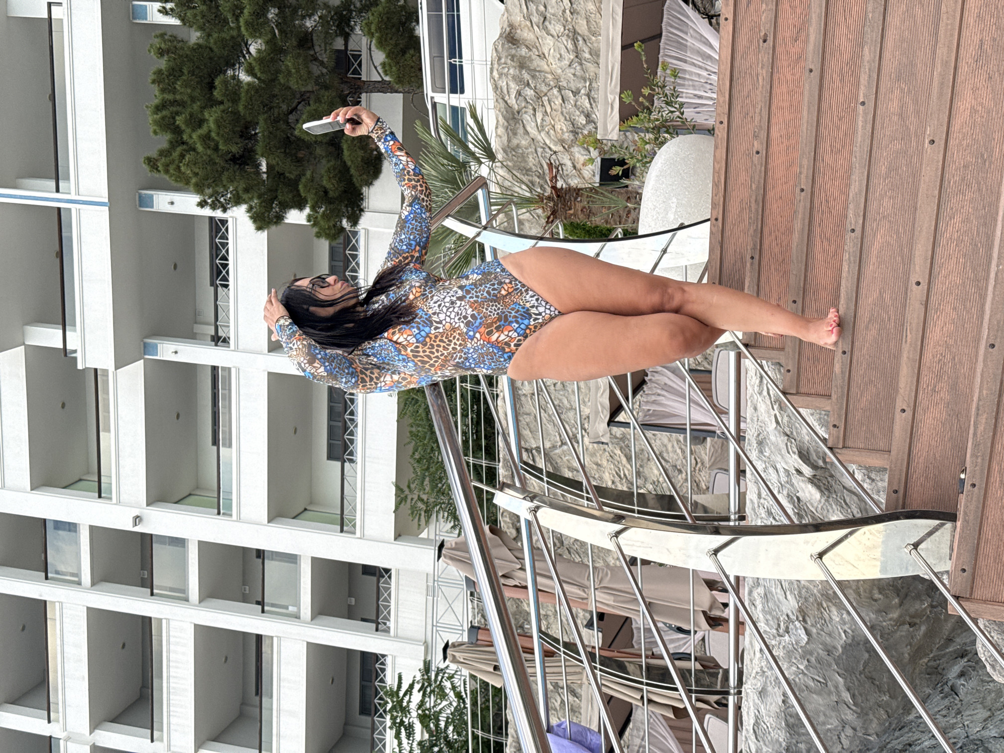 The image shows a woman in a long-sleeved swimsuit taking a selfie on a wooden deck next to a building. The woman has long, dark hair and is wearing glasses. Her swimsuit has a colorful, patterned design. She is holding a smartphone in her right hand, and her left hand is touching her head. Her legs are crossed, and she is looking at the phone. The building behind her is white with many balconies and windows. There is also a large green tree to the left of the woman. The setting appears to be a resort or hotel. The lighting suggests it is daytime, possibly late afternoon or early evening. The woman's posture and the selfie suggest a moment of leisure or self-expression. There is no visible text in the image.