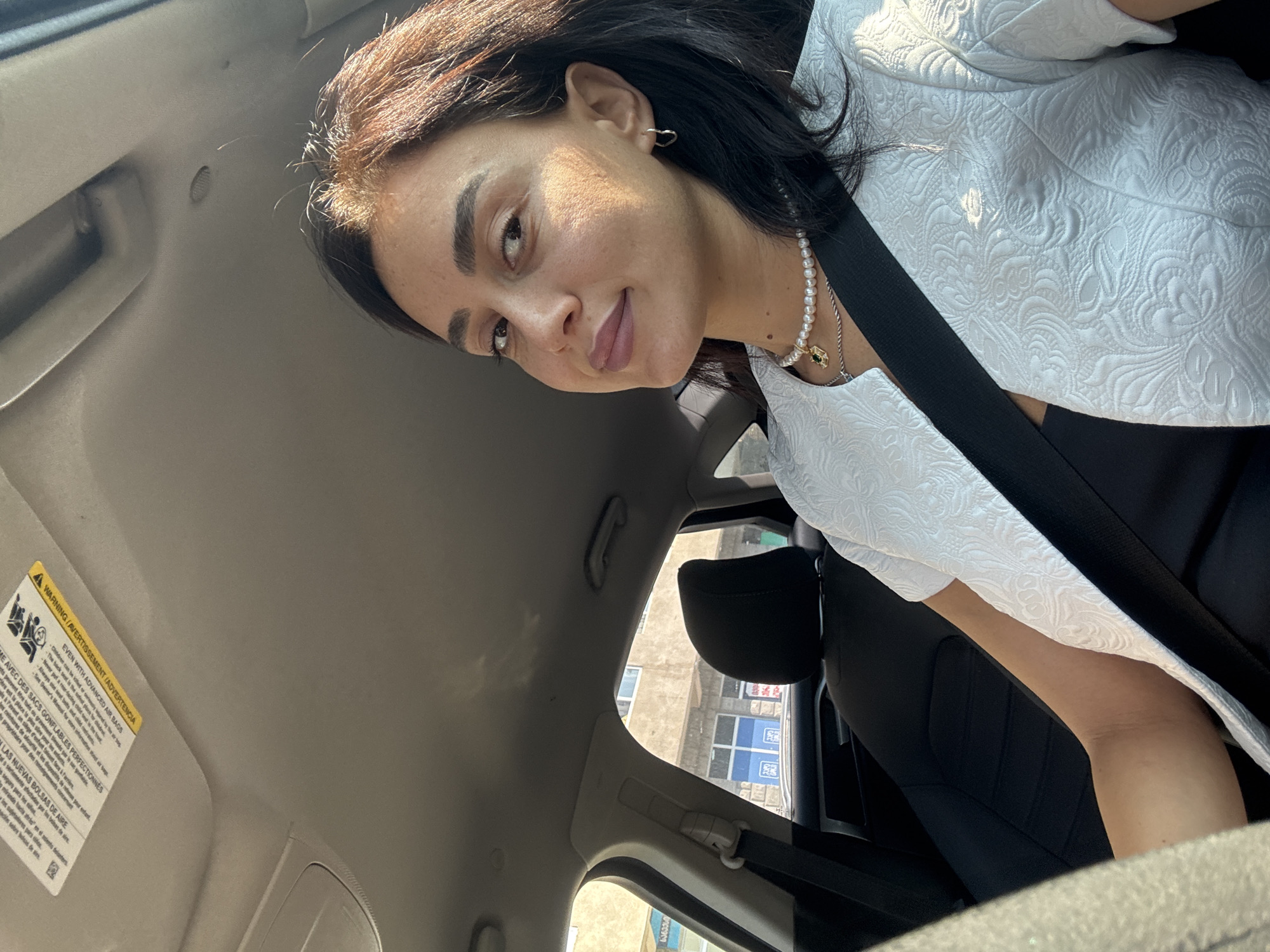 A woman with dark hair and fair skin is seated in the driver's seat of a car, looking towards the camera. She is wearing a white textured jacket over a black top and a pearl necklace with a green pendant. A black seatbelt is visible across her chest. The interior of the car is visible, including the headrest of the passenger seat. Sunlight streams in from the right, illuminating her face and jacket. Outside the car windows, buildings can be seen, suggesting an urban environment. Based on the text visible on a sticker on the sun visor, which appears to be in Georgian, the location is likely Tbilisi, Georgia.