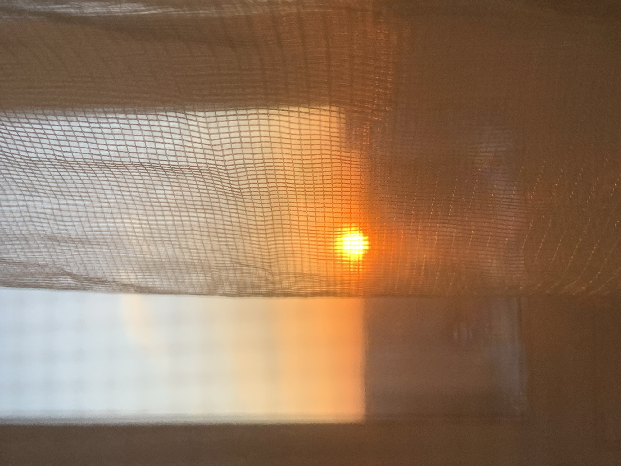 The image shows a close-up view through a sheer, possibly net-like curtain. A bright, circular light source, likely the sun, is visible through a gap in the curtain. The light casts a warm glow on the curtain fabric and partially illuminates the room beyond, revealing a blurred glimpse of a window and wall. The image does not provide information to determine that the photo was taken in Izhevsk, Russia.