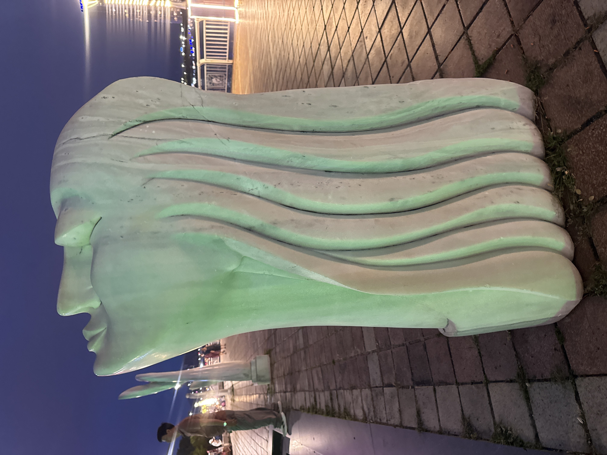 A medium-shot angled image depicts a stone sculpture of a human head with flowing hair. The sculpture is situated outdoors in what appears to be a public space, possibly a park or promenade. The background shows a waterfront scene with a building illuminated by lights, suggesting it may be evening. The environment around the sculpture is paved with stone blocks. A person is visible in the lower part of the frame near a similar sculpture, suggesting the presence of a series of such sculptures. Given the prompt context, this is probably taken in Đà Nẵng, Vietnam.