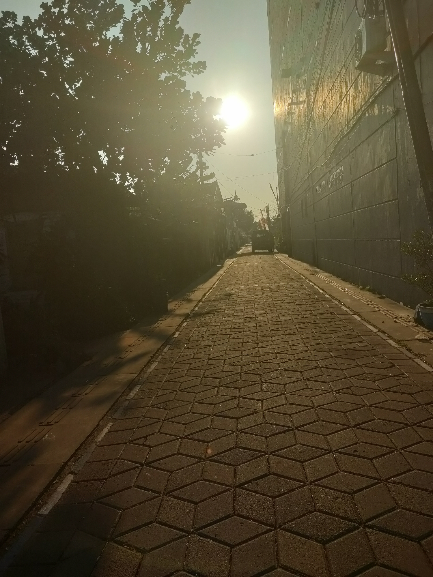 The image depicts a narrow, paved alleyway in Sooko, Indonesia, bathed in the warm light of a setting or rising sun. The sun's rays create a strong lens flare and cast long shadows, obscuring details on the left side of the image where a leafy tree and some buildings are visible. The right side of the alley is bordered by a tall, textured concrete building, likely an apartment complex or office building, with visible air conditioning units and electrical conduits. The alley floor is paved with hexagonal interlocking concrete blocks, arranged in a geometric pattern that creates a sense of depth. A small, dark-colored utility vehicle is parked in the distance, further down the alley. The overall impression is one of a quiet, urban residential or commercial area during a transitional time of day.