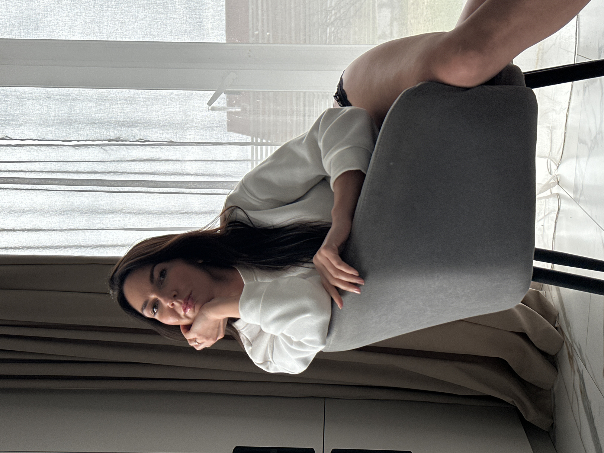 An indoor portrait captures a woman posing in a room with a large window. The woman is positioned on the right side of the frame, with her legs draped over the back of a contemporary grey upholstered chair, which has black metal legs. She is wearing a white long-sleeved top, and a glimpse of dark underwear is visible beneath her raised leg. Her long, dark hair flows over her shoulders and down her back.
Her upper body is angled towards the camera, and she rests her chin on her left hand, displaying a pensive or thoughtful expression with a slight pout. A ring is visible on her ring finger. Behind her, bright daylight filters through sheer white window blinds or curtains, creating a soft, diffused light. To the lower left of the frame, opaque brown or beige curtains are partially drawn, suggesting privacy or light control. The floor appears to be made of light-colored, possibly marble-patterned, tiles. Below the opaque curtains, a portion of white furniture or cabinetry is visible. The overall impression is one of a relaxed, contemplative moment indoors. There is no visible text in the image.