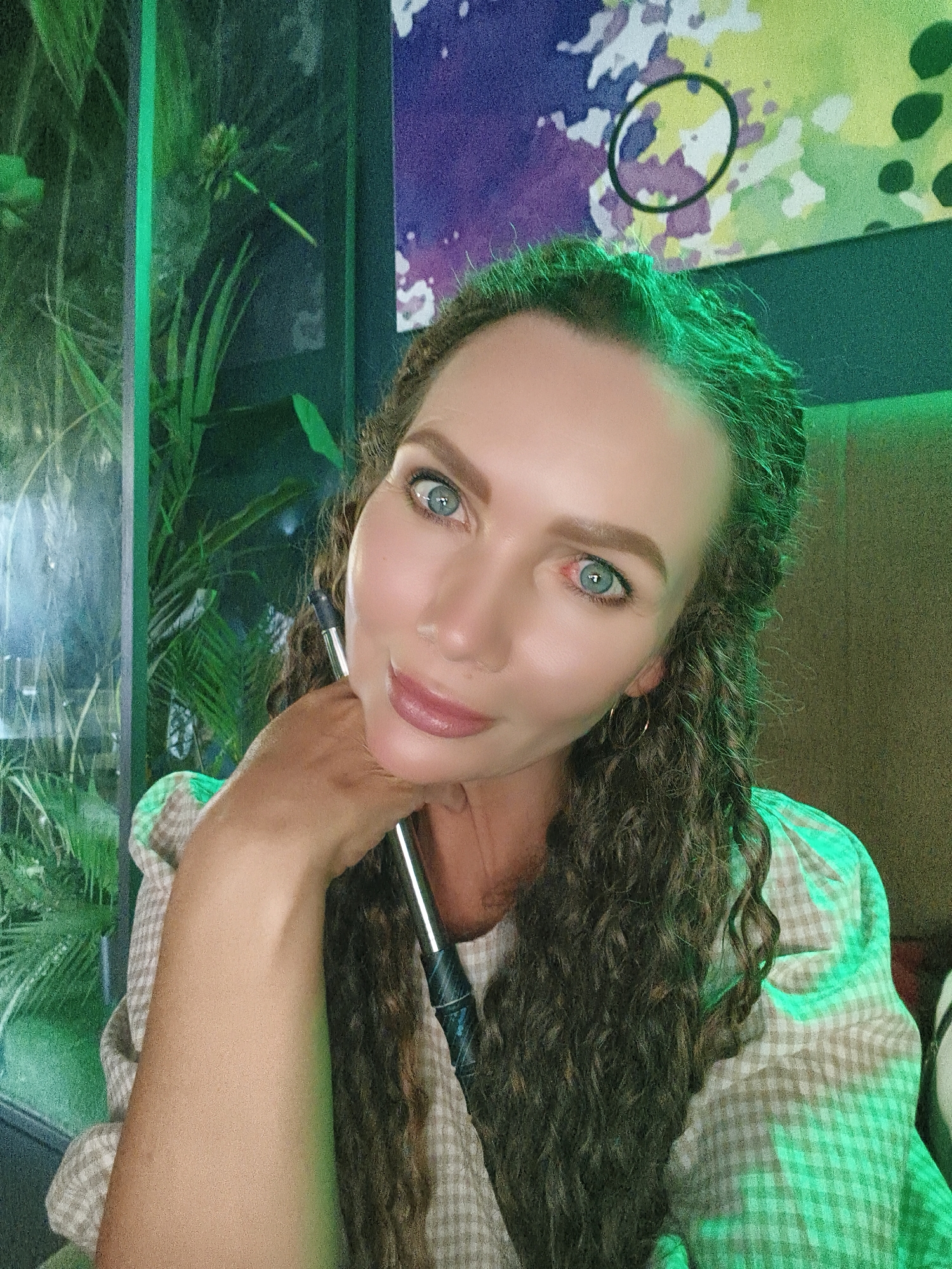 In an indoor setting with green ambient lighting, a woman with curly brown hair and blue eyes looks directly at the camera. She is resting her chin on her right hand, which holds a silver object. She wears a light-colored, checkered shirt. Behind her, lush green plants are visible through a glass enclosure on the left. The background on the right features a wall with an abstract, colorful mural and a textured surface. The overall scene appears to be a dimly lit lounge or restaurant.