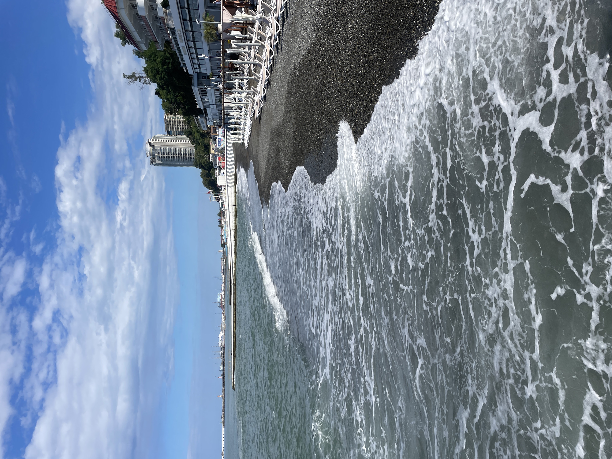 This image shows a view of a beach in Sochi, Russia, with the sea in the foreground and buildings and trees on the shore in the background. The sea is a dark, choppy blue-green with white foam from waves breaking on the shore. The beach is made of dark pebbles. Behind the beach, there is a promenade with rows of white lounge chairs and umbrellas. Further back, several buildings are visible, including a tall, modern high-rise building and a white multi-story hotel with a red roof. Trees are also visible on the hillside behind the buildings. In the distance, a port with cranes and ships can be seen, along with a lighthouse. The sky is blue with scattered white clouds.