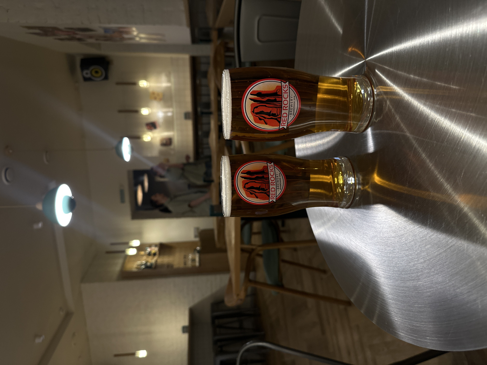 Here's a detailed description of the image:
Two full glasses of beer, with the "Red Rocks Brewery - Krasnoyarsk 2018" logo prominently displayed on each, sit on a brushed metal table. The scene is indoors, likely in a bar or restaurant. The background reveals a dimly lit interior with other tables and chairs, decorative lighting, and a mural or artwork on the wall. The orientation suggests that the photographer was likely sitting at the table.