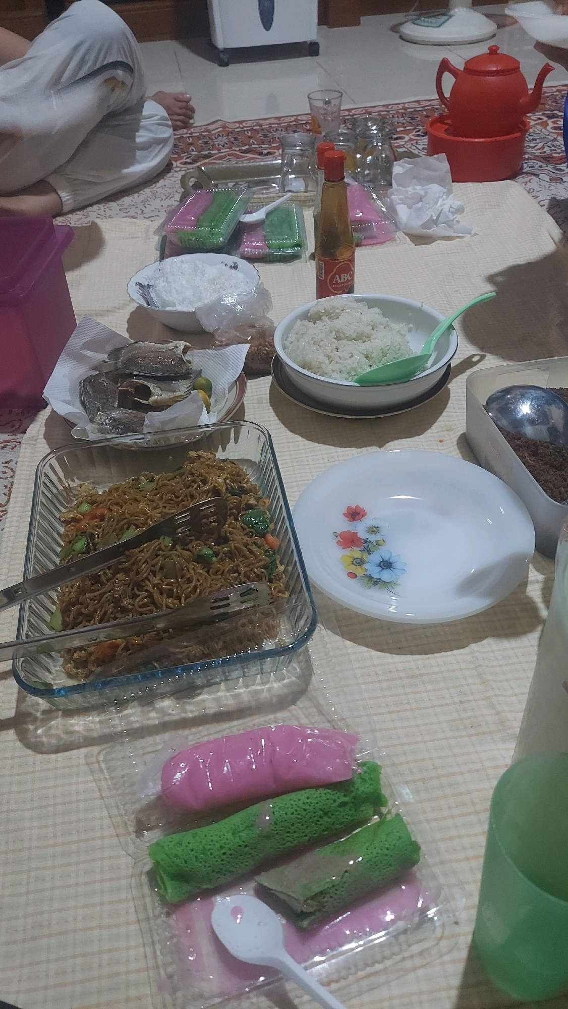 The image shows a casual meal laid out on a patterned mat. In the foreground, there are several sweet treats in a clear plastic container, including pink and green rolled pastries with a syrupy sauce. A white plastic spoon rests in the sauce. Behind the sweets, a glass baking dish contains what appears to be fried noodles with vegetables. Two metal serving tongs are placed in the noodles. To the right of the noodles is a white plate decorated with a floral pattern. Further back, there are bowls of white rice, a bottle of soy sauce, and a dish of dried fish with lime. In the background, a red teapot sits on a red base. Several glasses and jars are also visible. A person's legs, wearing white trousers, are visible on the left side of the image, suggesting they are seated on the floor. The overall scene depicts a home-style meal, possibly a gathering or family dinner, set on the floor. The presence of traditional Indonesian dishes like fried noodles, dried fish, and rolled sweets suggests the location might be Indonesia. Given the prompt requests a connection to Tanjung Redeb, Indonesia, this domestic scene is consistent with everyday life in such a location.