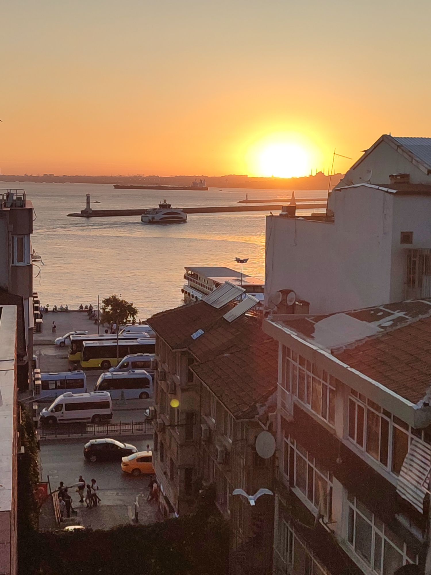 This image captures a sunset over the Sea of Marmara in Kadıköy, Turkey, as seen from a high vantage point. The sun, a bright orange orb, is low on the horizon, casting a warm glow across the water and sky. Silhouettes of buildings and a distant lighthouse are visible against the vibrant sunset. In the foreground, several buildings with tiled roofs dominate the view. Below, a street is lined with parked vehicles, including buses and cars. A group of people are gathered on a sidewalk, some walking and others standing. A ferry can be seen sailing on the water, and a seagull flies in the foreground. The overall scene depicts a bustling urban environment bathed in the soft, golden light of dusk.