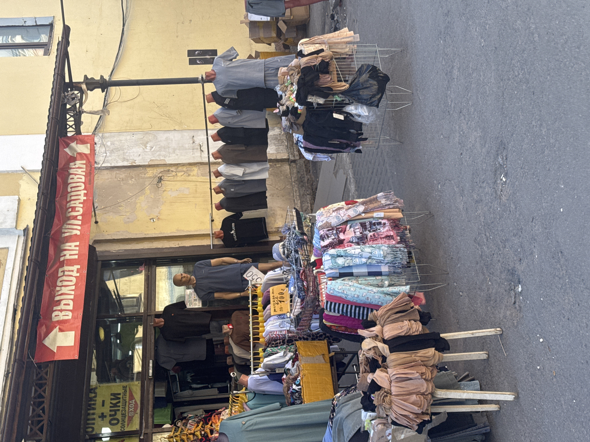 This media file depicts an outdoor market scene, likely in Saint Petersburg, Russia. The image is oriented vertically. In the foreground, racks of clothing are displayed for sale, including colorful scarves and folded garments. A large red banner with white text hangs from a building on the left, reading "ВЫХОД НА УЛ.САДОВАЯ" (Exit to Sadovaya Street) with an arrow pointing left. Below this, a yellow sign advertises "ОПТИКА" (Optics) and "СОЛНЦЕЗАЩИТНЫЕ ОЧКИ" (Sunglasses).
Further back, mannequins are dressed in t-shirts and pants, also displayed on racks. One mannequin appears to be wearing a blue t-shirt and shorts, with a price tag of "050" visible. Another mannequin is dressed in a black t-shirt with "RALPH LAUREN" text. The background shows a building with yellowing paint and some boxes stacked near an entrance. The ground is paved with asphalt. The lighting suggests it is daytime, with shadows indicating the sun is present. The overall impression is of a bustling street market where clothing and accessories are being sold.