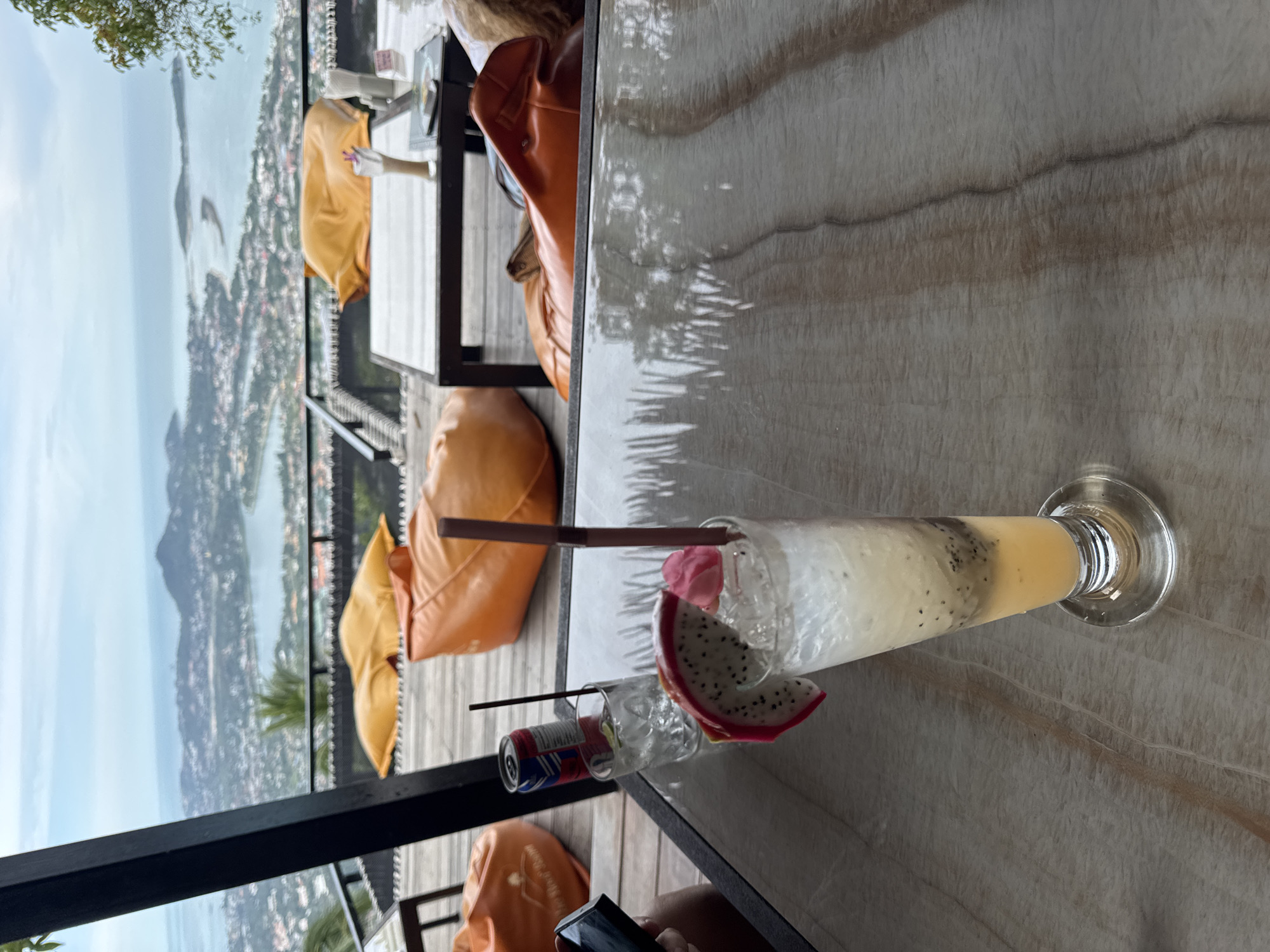 A tall glass of a fruity beverage with ice, a slice of dragon fruit, and a pink flower sits on a marble-topped table in the foreground. The drink is garnished with a dark brown straw and a small edible flower. Behind the table, a blurred landscape of a coastal city and the sea is visible under a cloudy sky. Several orange and yellow beanbag chairs are scattered on a wooden deck area, suggesting an outdoor seating area at a cafe or restaurant with a scenic view. A partially visible can of soda is also present on a nearby table. The overall scene depicts a relaxed outdoor dining experience with a breathtaking panoramic view.