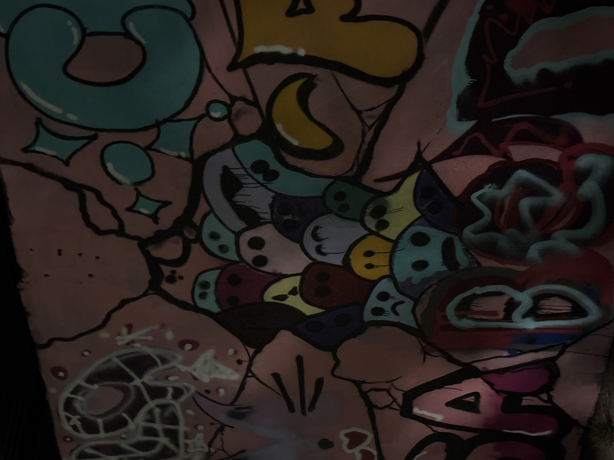 Graffiti covers a concrete wall. The artwork includes stylized letters, shapes, and cartoonish faces, rendered in various colors like teal, yellow, purple, and pink. There is a depiction of a rose. The wall appears to be outdoors, likely in a public space. Location is unknown.