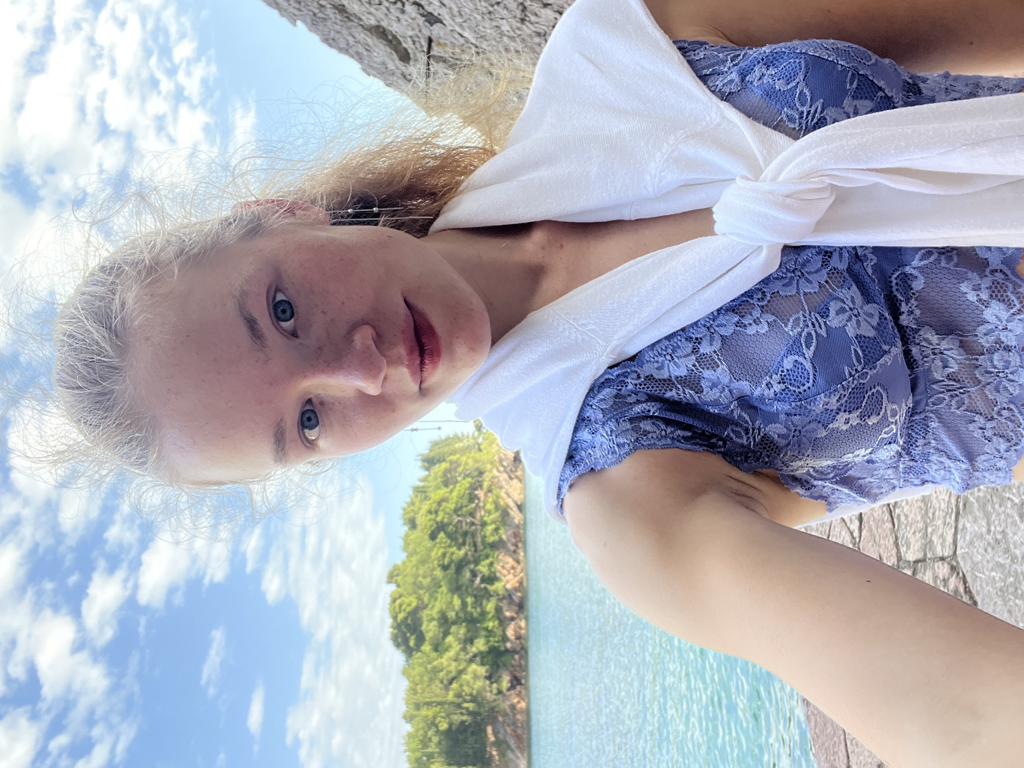 A young, fair-skinned woman with freckles and light blonde hair is the subject of this selfie, with the background providing a hint of the location as Rađenovići, Montenegro. She has blue eyes and is wearing a light blue lace top, over which a white garment is draped and knotted at the center. The background shows a sunny sky with scattered clouds, turquoise water, and a green, tree-covered island. A stone structure is also visible in the frame. The orientation of the image is sideways.