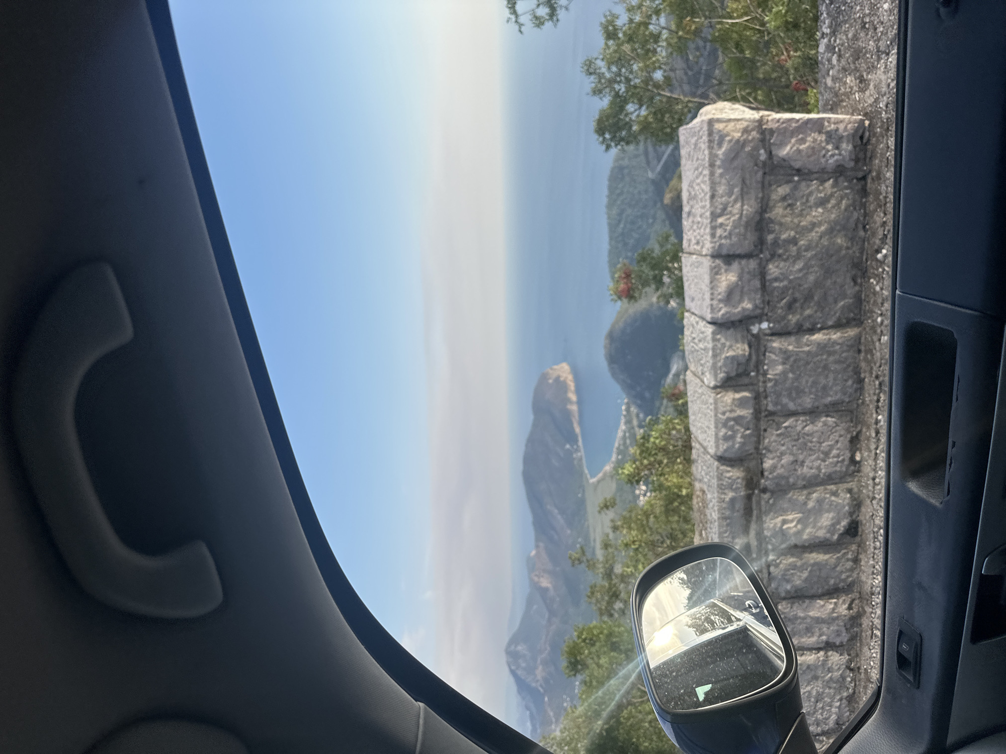 The image, taken from inside a vehicle, captures a scenic view of the coast and mountains near Novoselje, Montenegro. The foreground shows the interior of the car, including a portion of the dashboard and door panel, along with a side mirror reflecting the bright sun. A rough stone wall lines the edge of the road, with green foliage growing on either side.
Beyond the wall, a dramatic landscape unfolds. Rolling green hills and mountains stretch towards the sea, with a bay visible in the distance. The water is a deep blue, meeting the horizon under a clear, pale blue sky. The overall impression is one of natural beauty and a peaceful, expansive vista.