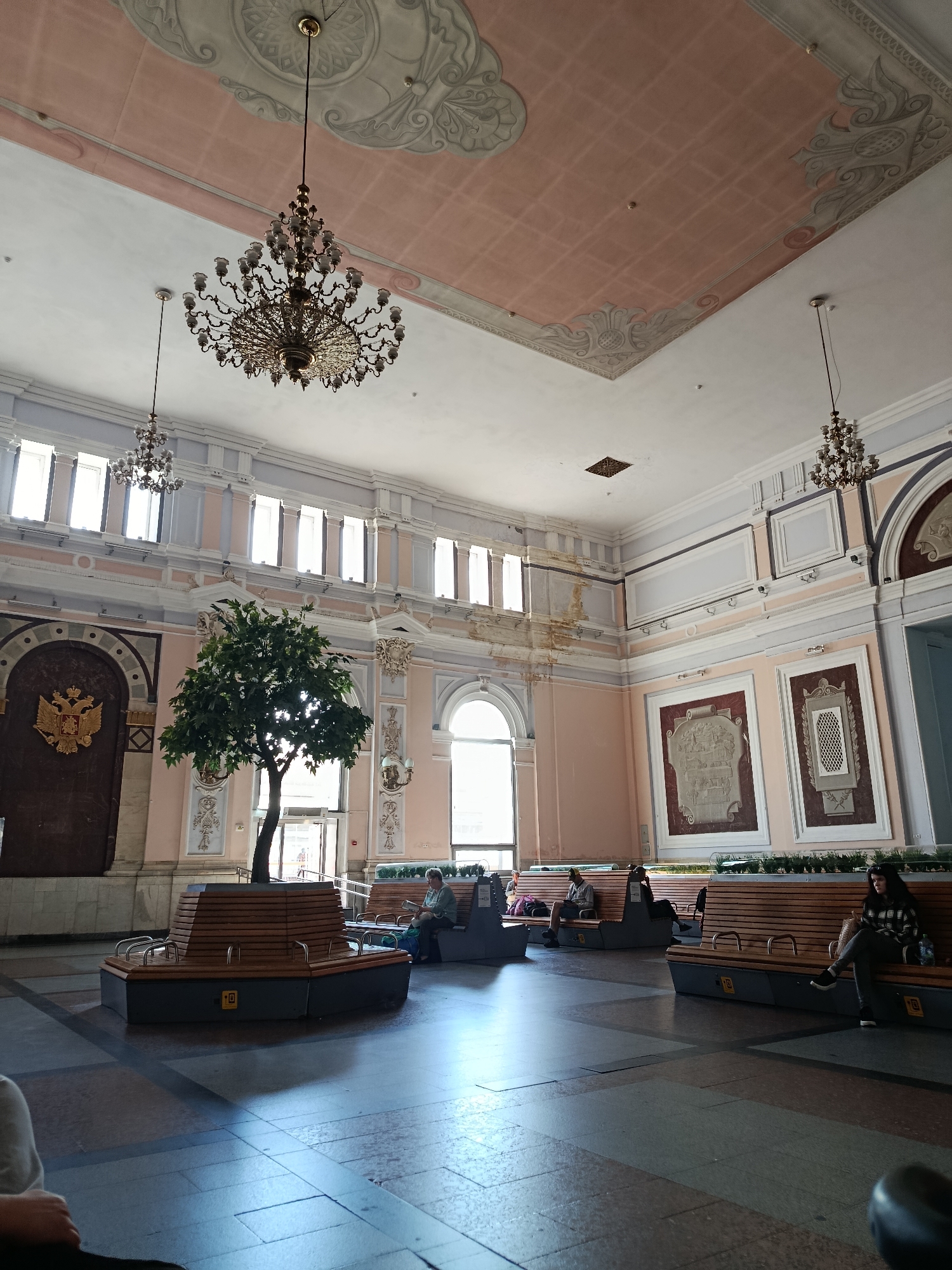 The image depicts the interior of a grand waiting hall, likely a train station, with high ceilings adorned with ornate stucco work and large chandeliers. The walls are painted in soft pink and pastel blue hues, with decorative panels and large windows that allow natural light to stream in. In the center of the hall, a large tree is planted within a wooden seating area, surrounded by benches where several people are seated, appearing to be waiting or resting. The floor is tiled in a geometric pattern of dark and light gray squares.
The building's architecture suggests a historical significance, possibly dating back to the late 19th or early 20th century, with elements reminiscent of Beaux-Arts or Art Nouveau styles. The presence of a large imperial crest on the left wall, featuring a double-headed eagle, strongly indicates that this location is in Russia, and potentially Moscow, given the grandeur.
Specifically, this appears to be the waiting hall of Yaroslavsky Rail Terminal (Yaroslavsky Station) in Moscow, Russia. The distinctive architectural features, including the ceiling ornamentation, the chandeliers, the layout of the waiting area with benches and the central tree, and the prominent imperial crest, are all characteristic of this historic Moscow landmark.
There are at least five individuals visible in the image, all seated on benches. One woman in the foreground right is wearing a checkered jacket and jeans, looking down at her belongings. Another person is sitting further down the same bench, mostly obscured. To the left, in the mid-ground, a few more individuals are seated on other benches, some reading or looking ahead. The atmosphere is calm and quiet, with a sense of anticipation.