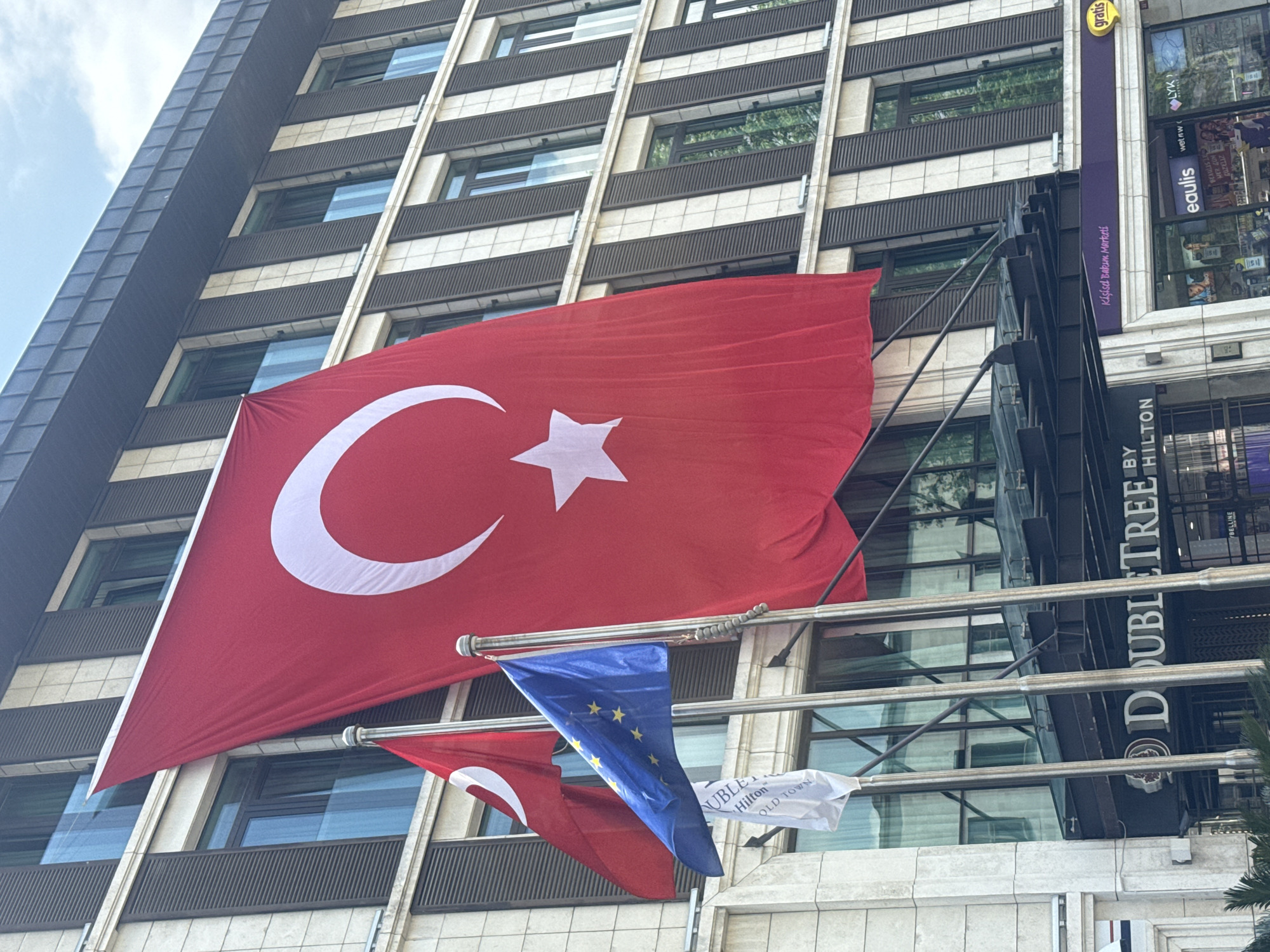 Here is a detailed description of the image:
**Environment:** The image presents an urban outdoor scene. The primary focal point is a tall building with a repetitive pattern of windows. The sky is visible in the upper left corner, suggesting daytime.
**Objects:**
- Prominently displayed is the flag of Turkey, red with a white crescent moon and star.
- The flag of the European Union, blue with yellow stars, hangs just below the Turkish flag.
- A smaller white flag with the text "DOUBLETREE BY HILTON OLD TOWN" is also present.
- Signage for "gratis" and "eaulis" is visible, indicating commercial establishments within the building.
- The “DOUBLETREE BY HILTON” branding is prominently displayed on the building's facade.
**Subjects and Interactions:**
- There are no people visible in the frame. The flags and branding suggest this location is a hotel with a commercial element.
**Location:** This is likely the DoubleTree by Hilton Istanbul-Old Town. The Turkish flag is prominently displayed along with EU and hotel branding. The commercial shops inside confirm an urban, commercial setting.