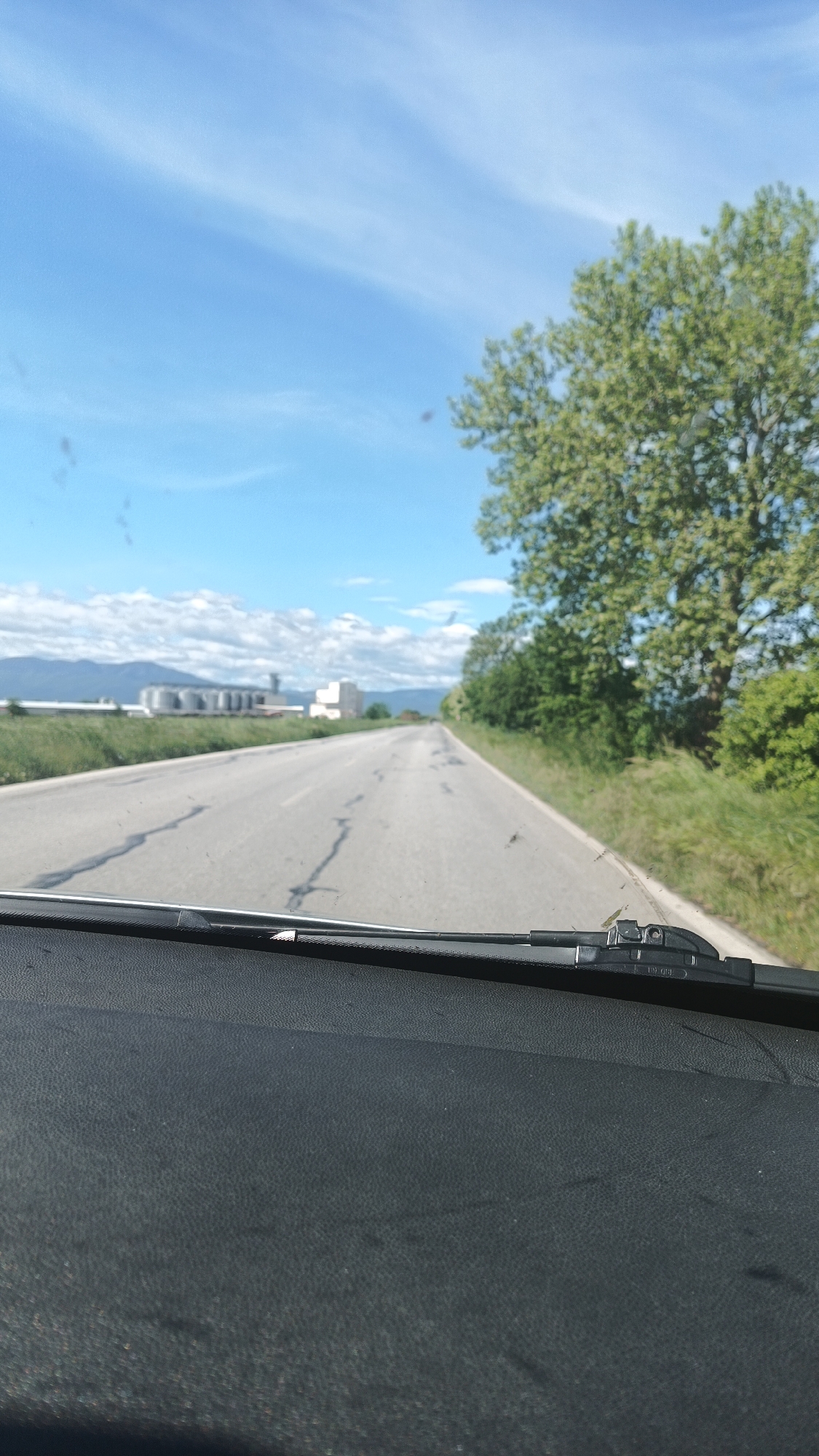 The image shows a view from inside a vehicle, likely a car, looking out onto a straight, cracked asphalt road. The road is flanked by grass and trees on the right side. In the distance, on the left side of the road, there appears to be a large industrial complex with silos and buildings. The sky is blue with scattered clouds. The dashboard of the vehicle is visible in the foreground, including the windshield wipers. The location is inferred to be near Bogdanitsa, Bulgaria, based on the presence of agricultural or industrial structures and the flat, rural landscape. There is no indication of people in the image.
