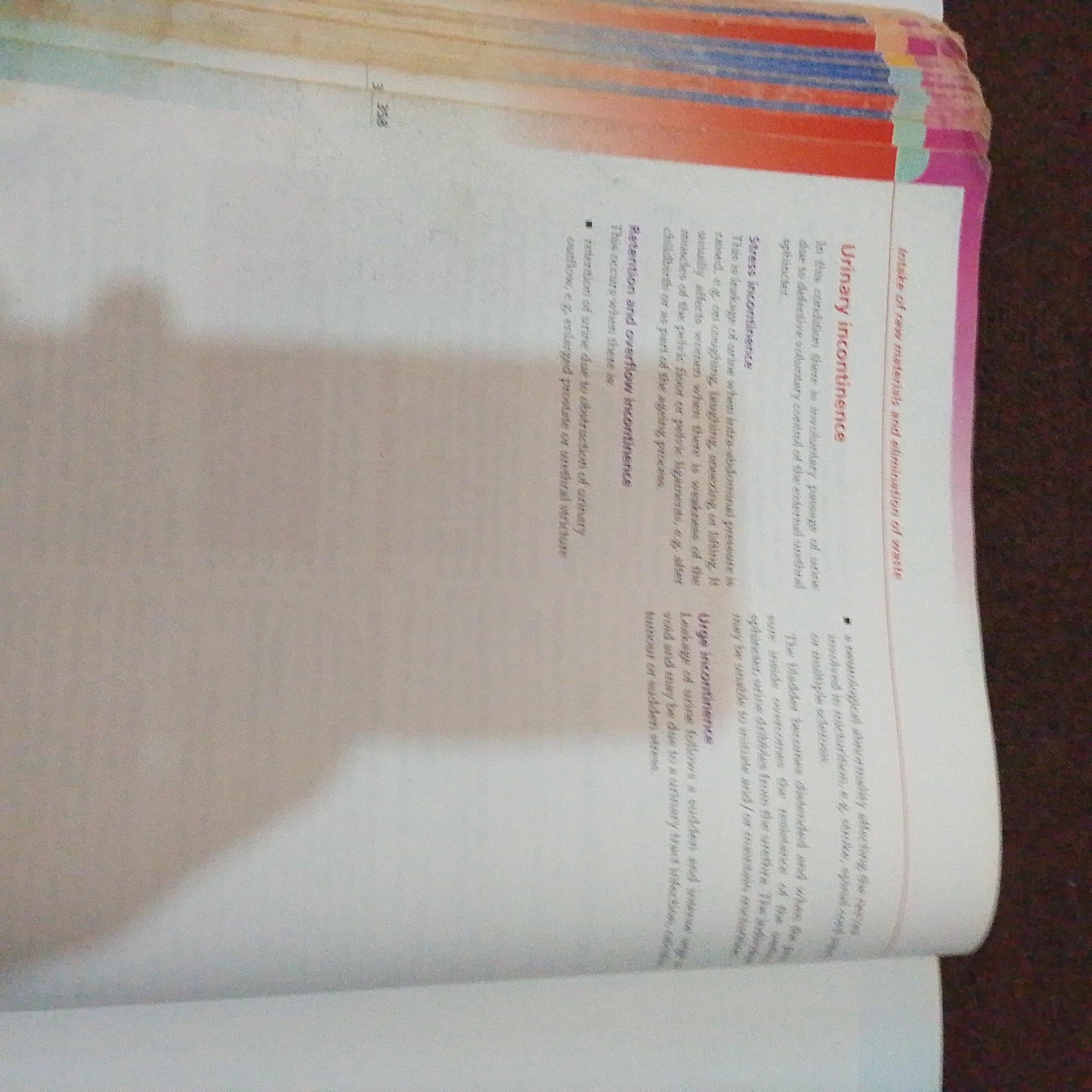 The image displays a page from a textbook. The visible text pertains to urinary incontinence and its different types: stress incontinence, retention and overflow incontinence, and urge incontinence. The page also includes a heading that reads "Urinary incontinence" and another that reads "Intake of raw materials and elimination of waste." There are also bullet points and descriptive paragraphs explaining these conditions. The physical context of the image is a book lying open on a surface, with some of the binding and pages visible on the right side, appearing slightly worn. The lighting suggests an indoor setting. There are no people or any elements that would indicate a location in Jalingo, Nigeria.