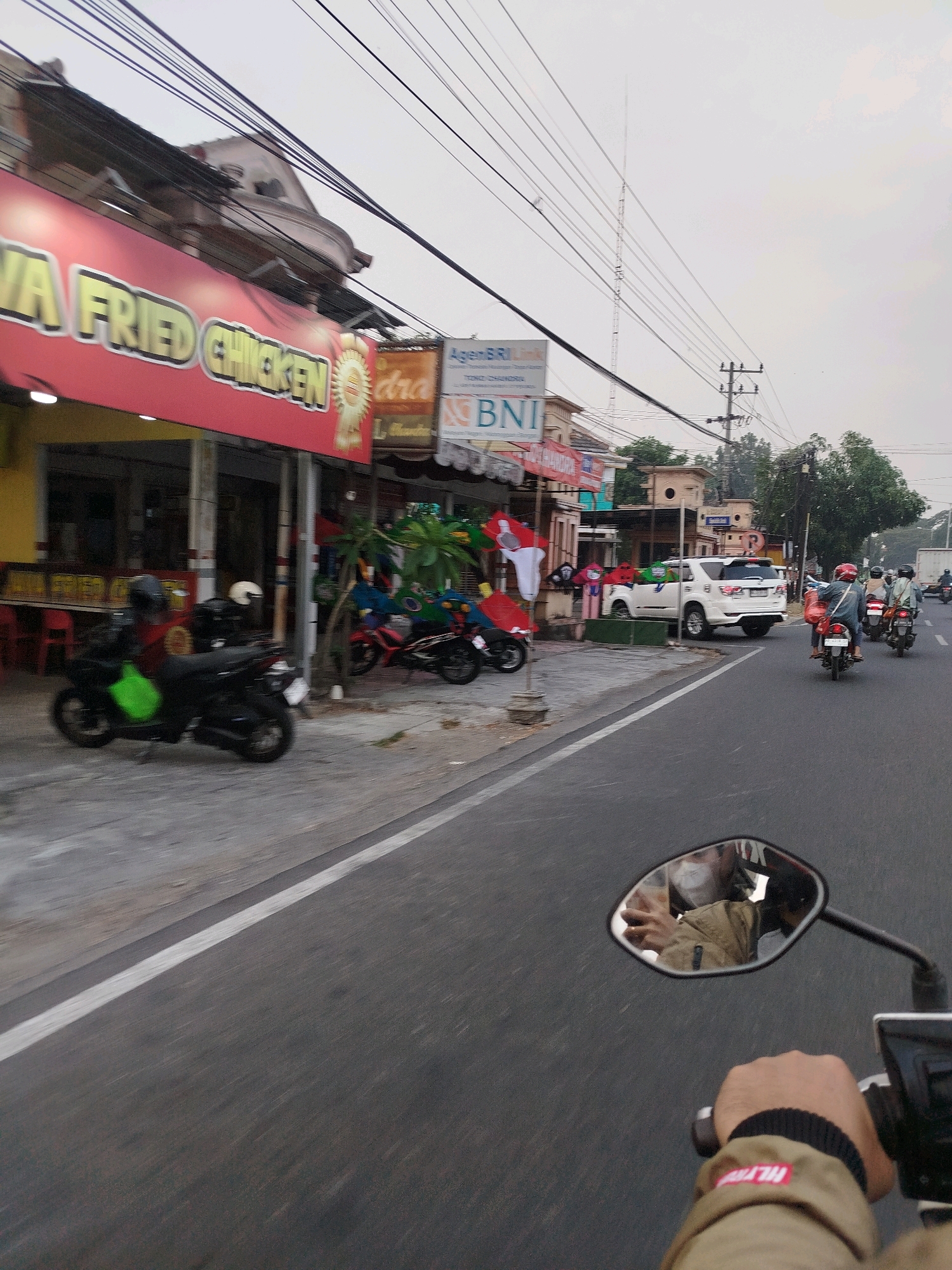The image captures a street scene in Cekok, Indonesia, with a clear focus on traffic and commercial establishments. A motorcycle, ridden by a person wearing a tan jacket and a black beanie, is in the foreground, with its handlebar and a side-view mirror visible. The mirror reflects the rider's face, partially obscured by a mask, and the surrounding environment. A white car is parked on the side of the road, alongside other motorcycles. A prominent red and yellow sign for "JAVA FRIED CHICKEN" hangs above a storefront, indicating a food establishment. In the background, utility poles and wires crisscross the sky, and a tall tower is visible in the distance. The overall scene suggests a typical urban environment with ongoing traffic and daily commerce.