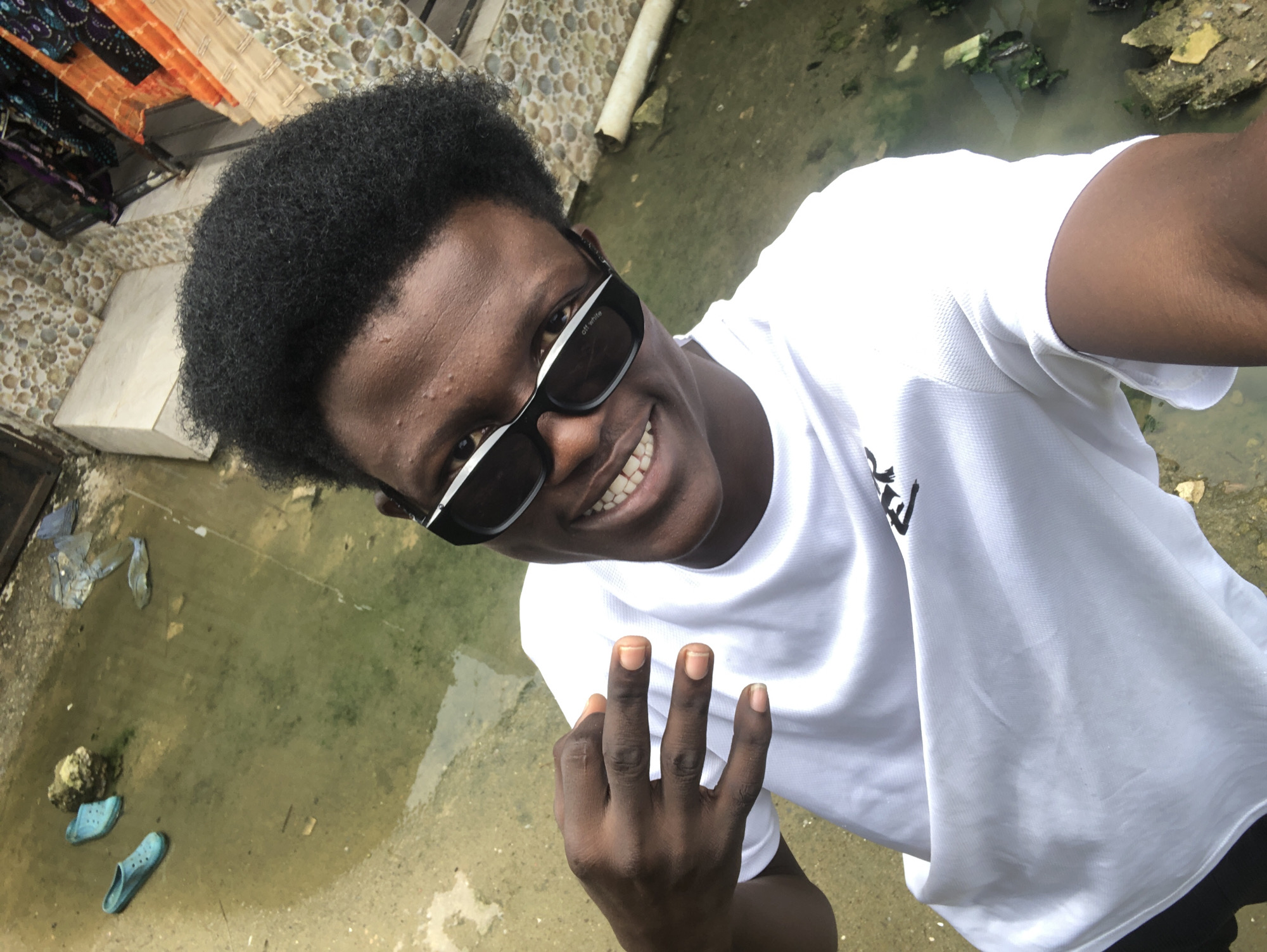 A young Black man with dark skin and short, curly black hair, wearing black-rimmed sunglasses and a white t-shirt, poses for a selfie. He is smiling broadly, showing his teeth. His left hand is raised, with his index, middle, and ring fingers extended, and his thumb slightly bent. He is positioned in the foreground, with the background showing a murky, shallow pool of water on a concrete surface. In the water, to the left, are two blue slip-on shoes. The background also features a textured wall with small, colorful mosaic-like tiles, and some hanging fabrics in orange and purple. The overall environment appears to be an outdoor, potentially urban, setting, possibly a courtyard or back alley, and the presence of the shoes and water suggests a less maintained area. This scene is located in Ado, Nigeria.
