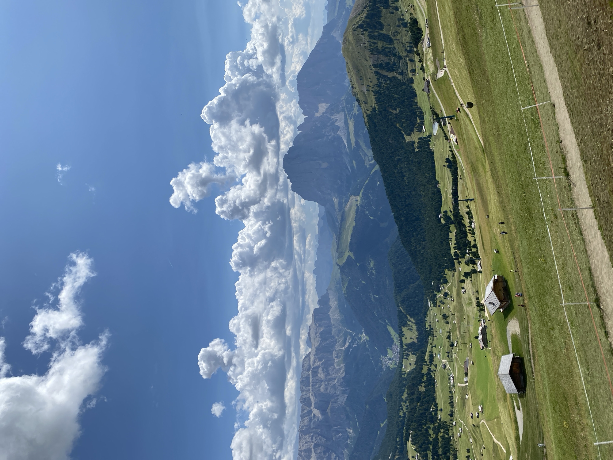 The image depicts a panoramic view of a mountain landscape in the Dolomites, Italy. The foreground features rolling green hills dotted with small wooden buildings, possibly chalets or farmhouses. A dirt path winds through the grassy slopes, with several people walking along it. Further in the background, dense green forests cover the lower slopes of the mountains. Towering above are rugged, majestic mountain peaks, their rocky formations stark against a bright blue sky filled with fluffy white clouds. The scene suggests a sunny day in a mountainous region, likely a popular tourist destination for hiking and outdoor activities. The overall impression is one of natural beauty and tranquility.