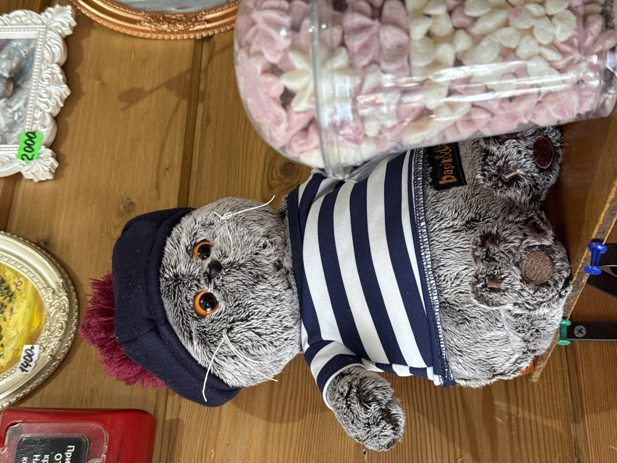 On a wooden surface, a stuffed animal cat with orange eyes wears a dark blue striped shirt and a matching hat with a maroon pom-pom. A clear jar filled with pink and white meringue candies partially obscures the cat's body. To the left are two framed items with price tags of 2000 and 1400. A red object with a partial label in Russian is at the bottom left. The location is unknown.