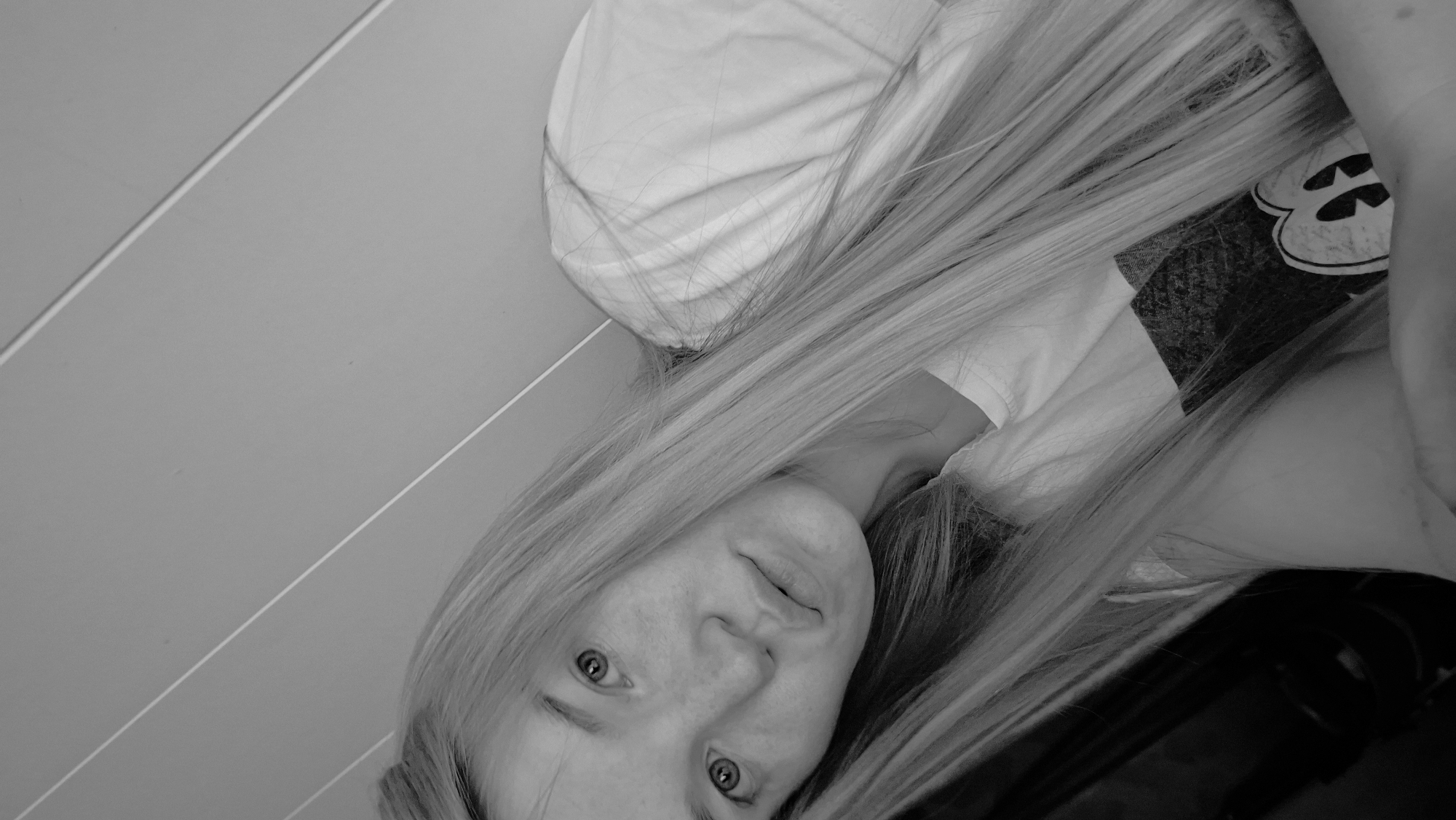 A black and white, close-up, head and shoulders selfie of a young Caucasian woman with long, straight blonde hair is captured. She is wearing a t-shirt with a graphic of Mickey Mouse on it. The background consists of a slanted wall with three thin, parallel lines. The woman is looking directly at the camera with her head tilted. The lighting is soft, casting a shadow on the right side of her face. There is no discernible information about the location in the city of Большой Букор, Russia.
