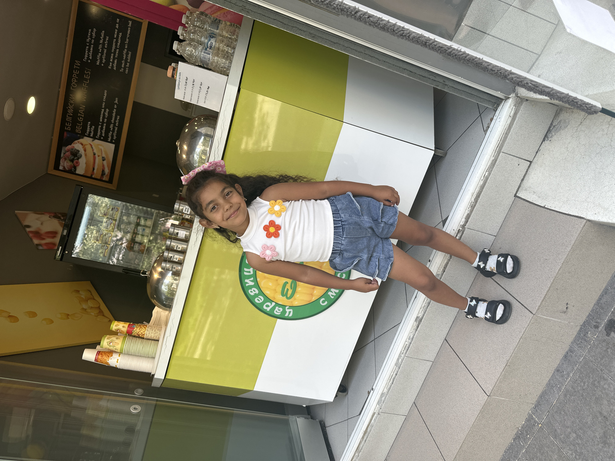 A young girl with dark curly hair and a pink bow stands on a tiled outdoor staircase in front of a yellow and white food stand. She is wearing a white sleeveless top decorated with floral appliques, denim shorts, white socks with butterfly designs, and black platform sandals. The food stand has a menu board displaying "BELGIAN WAFFLES" in English and Bulgarian, and a selection of cups and bottles are visible. The girl is smiling at the camera and her left hand is slightly bent at the elbow. The environment appears to be a street or walkway in a city.