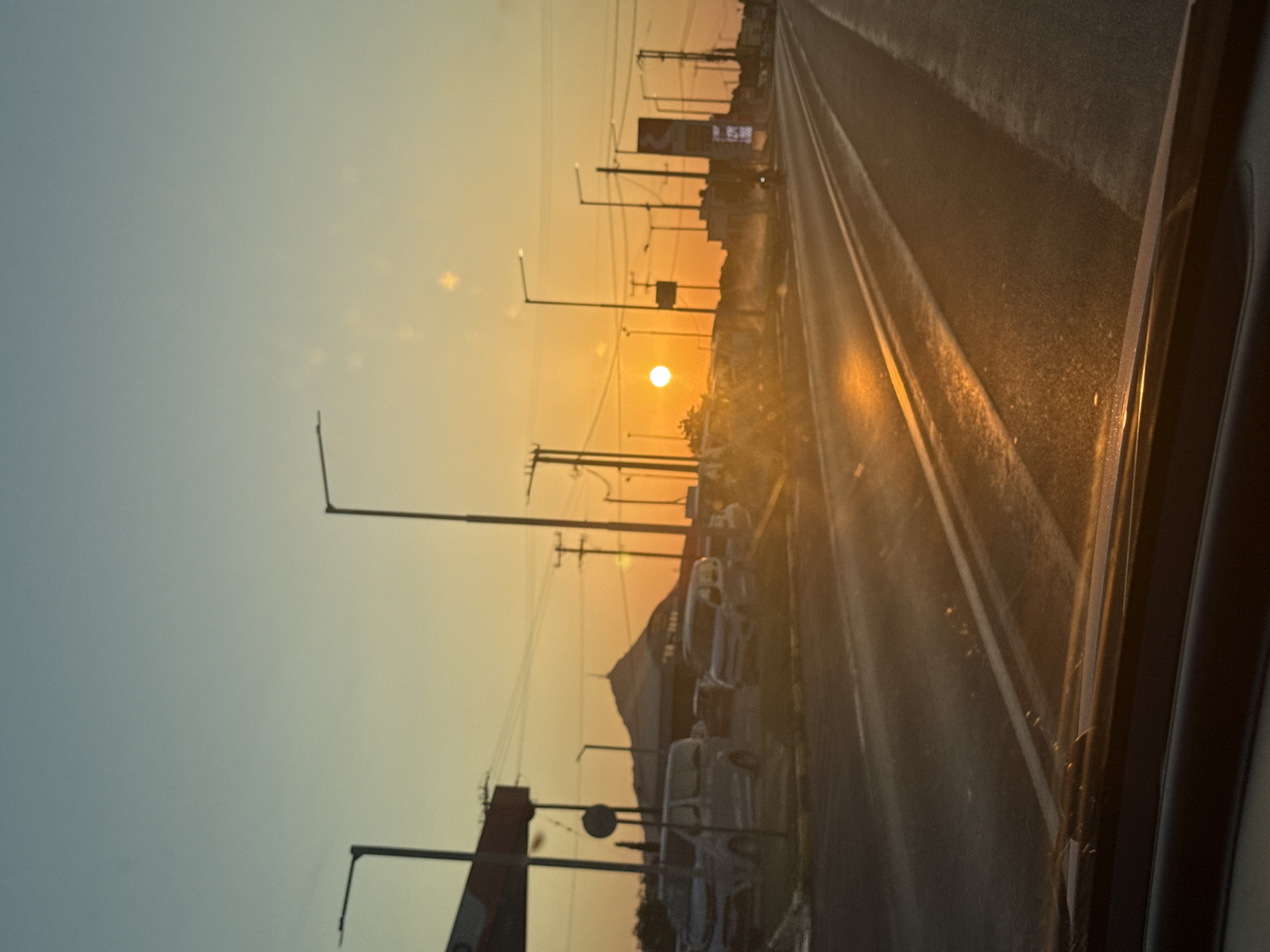 A street-level, eye-level, horizontal, medium shot captures a sunset scene on a road. The sun, a bright orange orb, is setting on the horizon, casting a warm glow across the sky and the road. The sky transitions from a pale blue at the top to a vibrant orange near the horizon. Several streetlights are visible along the side of the road, their tall poles reaching towards the sky. A line of parked vehicles, including vans and cars, is visible on the left side of the road. In the background, a domed building stands prominently, silhouetted against the sunset. A tall, illuminated sign is also visible on the right side of the road. The road itself appears wet, reflecting the orange light of the setting sun. The foreground shows the lower edge of a car's windshield and dashboard, suggesting the photograph was taken from inside a vehicle. The overall atmosphere is serene and tranquil, with the setting sun creating a picturesque view of an urban landscape.
