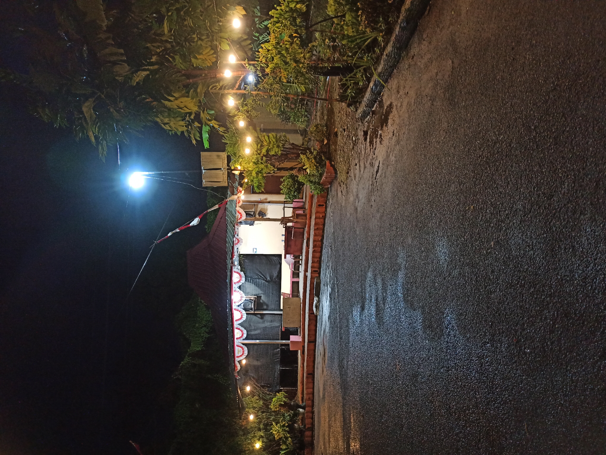 The image depicts an outdoor scene at night, likely in a public space such as a park or a small food stall area, located in the City of Medan, Indonesia. A modest, open-air structure with a red, gabled roof is illuminated by string lights and a bright overhead lamp. The structure appears to be a food stall or a small building with a covered area. Red and white bunting decorates the front of the structure.
In front of the structure, there are several tables and chairs, suggesting it is a seating area. The ground in front of this area is paved and appears wet, reflecting the lights. To the right of the structure, there are trees and dense foliage, also illuminated by string lights. A tall pole with a bright light is visible in the background, casting a strong glow. The overall impression is one of a simple, perhaps family-run, establishment operating in the evening. The presence of Indonesian flags (red and white) on the structure indicates a possible celebration or patriotic display.